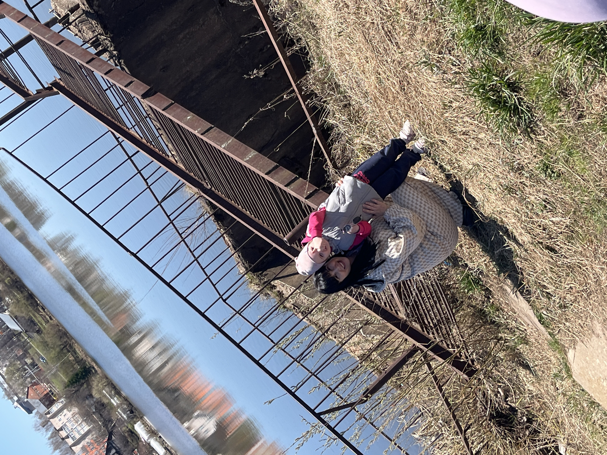 The image captures two individuals, a younger person on top and an older person, presumably a parent, below, both lying on a platform composed of brown metal grating. This platform is partially suspended over what appears to be a body of water reflecting the sky and buildings, indicative of a bridge-like structure. The environment includes dry grass and vegetation adjacent to the metal structure. The location is Zav'yalovo, Russia, based on information provided.