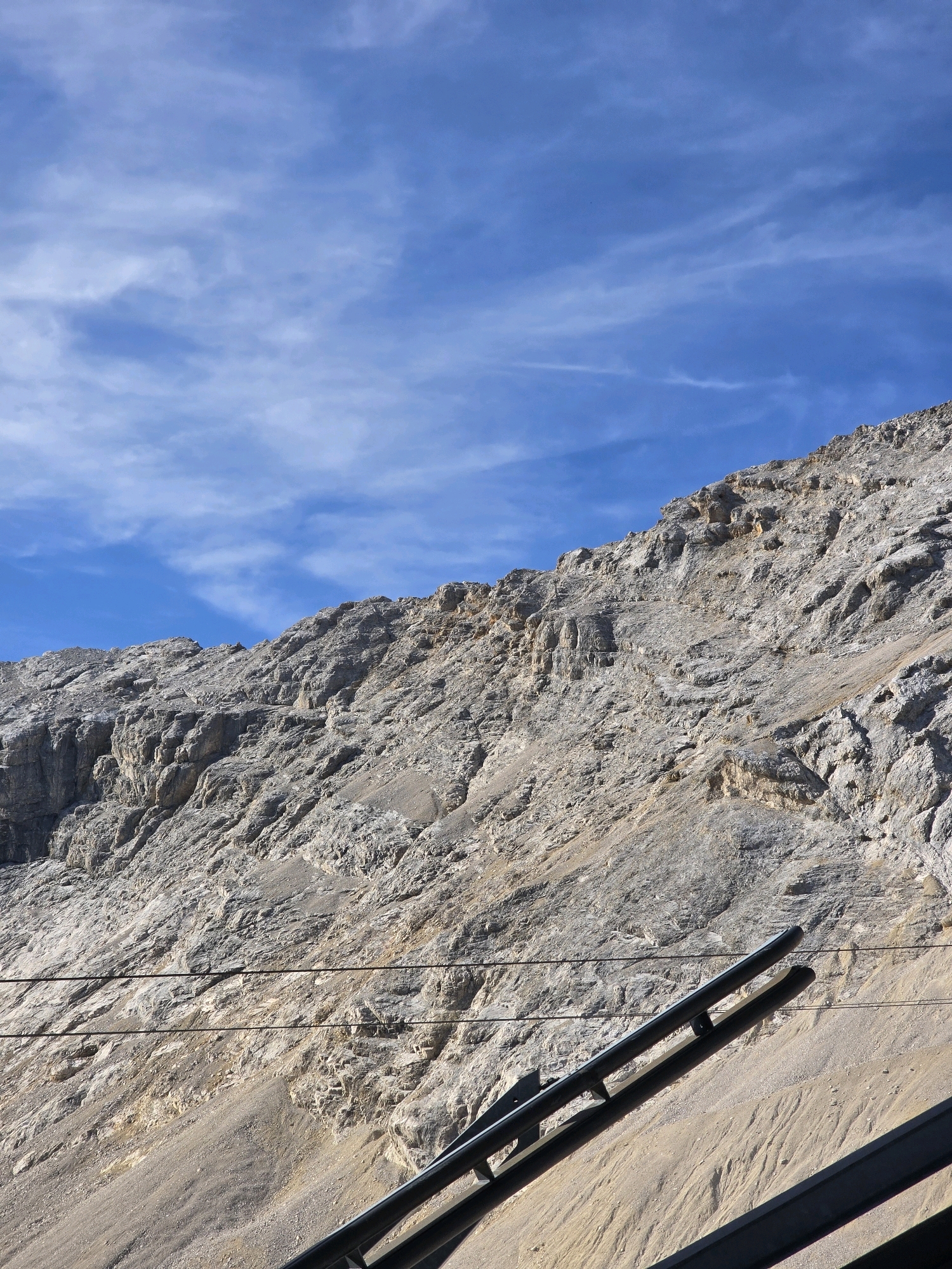 This image shows a steep, rocky mountainside under a bright blue sky with wispy white clouds. The mountain is textured with various shades of grey and brown rock formations, suggesting rugged terrain. In the foreground, a dark metal railing and support structure is visible, angled upwards, indicating it might be part of a viewing platform, a cable car station, or a hiking trail infrastructure. Two parallel dark cables run horizontally across the lower half of the image, further suggesting the presence of a cable car system. This scene is consistent with the Zugspitze, Germany's highest peak, located near Garmisch-Partenkirchen. There are no people visible in the image.