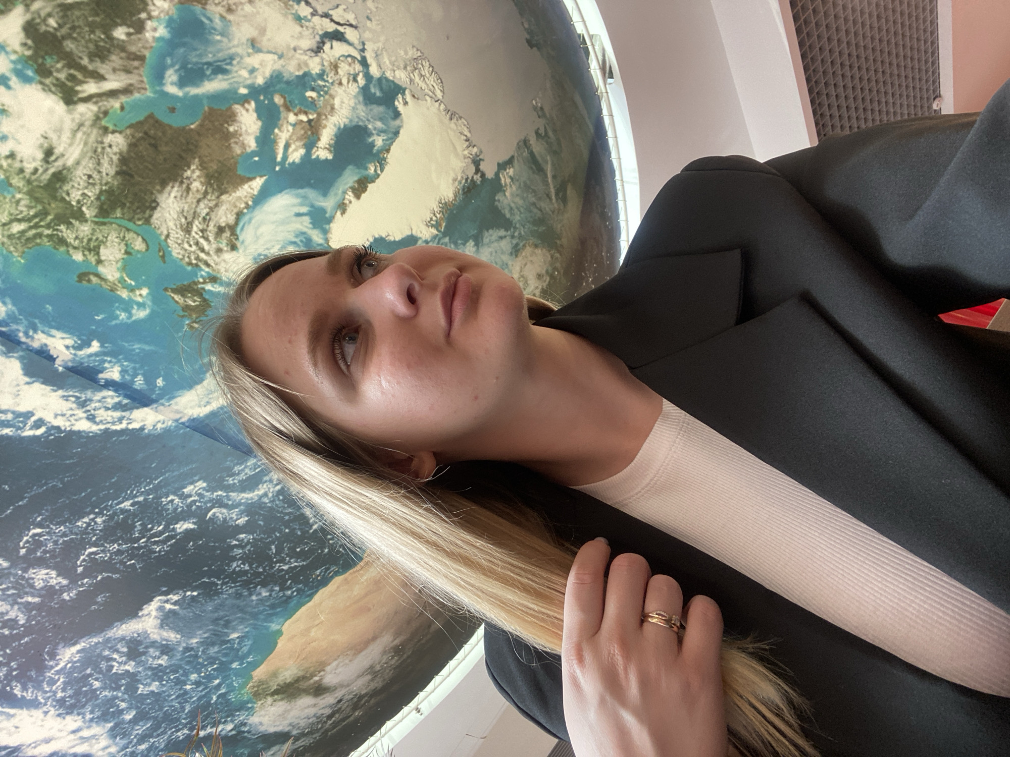 The image shows a young woman with blonde hair and fair skin posing for a selfie. She is wearing a white ribbed shirt and a black blazer. Her left hand, adorned with two rings, is touching her hair. In the background, a large, spherical map or model of the Earth is visible, with clouds and landmasses depicted. The lighting suggests an indoor setting, possibly an educational institution or museum, given the Earth model. The environment appears to be well-lit, with a ceiling structure visible in the upper right corner. There are no discernible landmarks or street views to pinpoint a specific location within Izhevsk, Russia. The image is a close-up shot, focusing on the woman and the immediate background.
