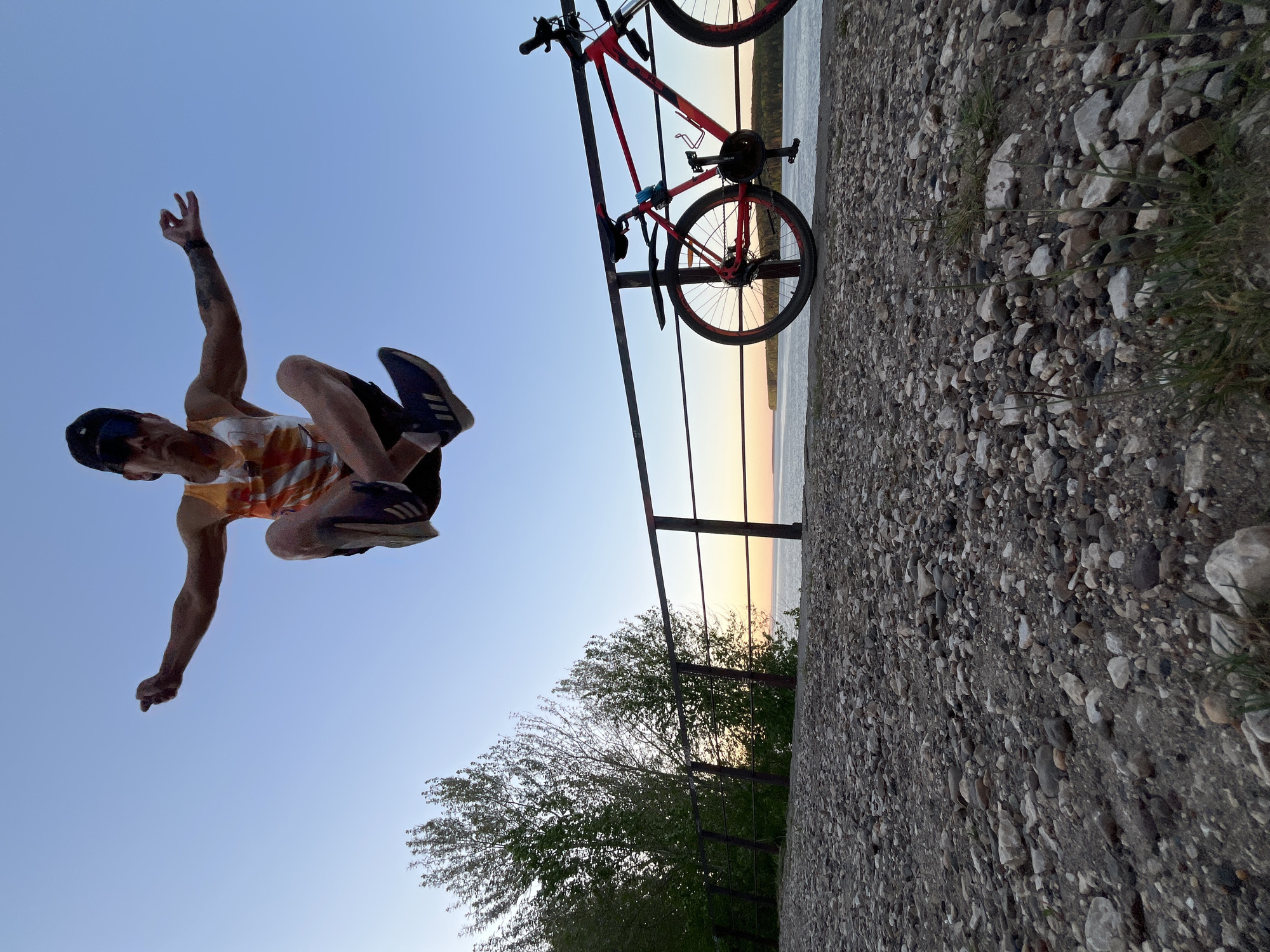 A man wearing a multicolored tank top, black shorts, and dark sneakers appears to be suspended in the air with his arms extended, photographed from a low angle. A red bicycle leans against a metal railing in the background. The railing separates a rocky terrain from a body of water reflecting the sunset. Trees line the edge of the bank. The sky is clear. The photograph was taken near the city of Chaikovsky, Russia.