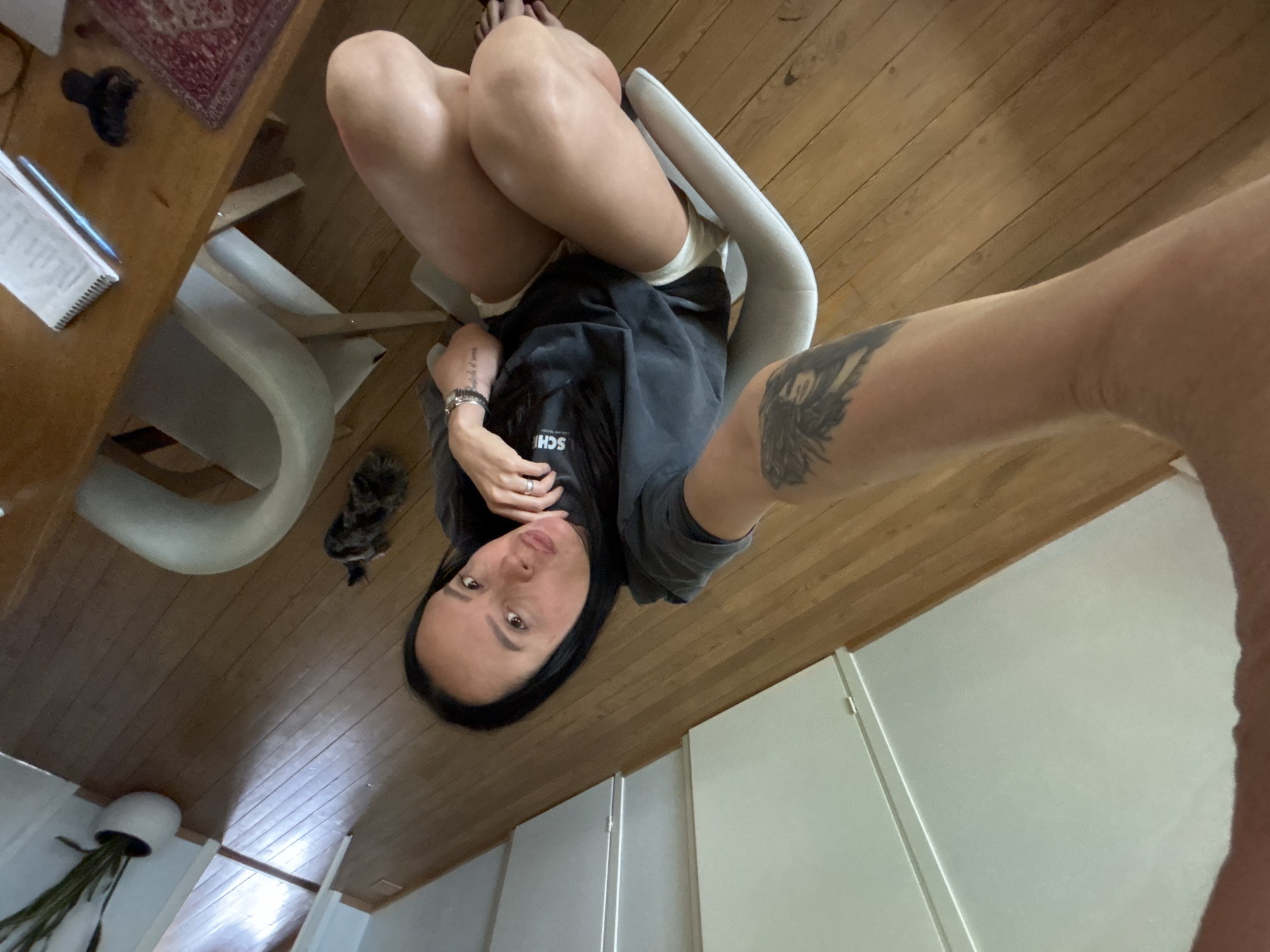 A person is sitting in a white chair in a room with wood floors. The person has dark hair and is wearing a grey sweatshirt. Their legs are crossed. A table with a notebook and other items is visible in the background. A white cabinet and a plant are also visible. The image appears to be a selfie. There is no information to confirm this image was taken in Chaikovsky, Russia.