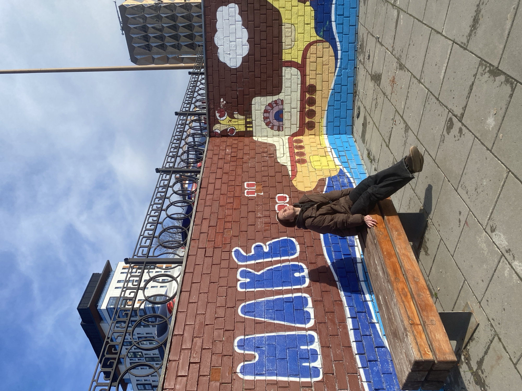 A woman in a brown jacket and dark pants lies on a wooden bench next to a brick wall painted with a mural, which is located in Yekaterinburg, Russia. The mural has a blue and white word painted on it, and above that is a drawing of a beige and brown object resembling a telephone or communication device. Above the mural is a decorative black metal fence. In the background, buildings and the sky are visible. The bench sits on a paved area with gray rectangular stones.