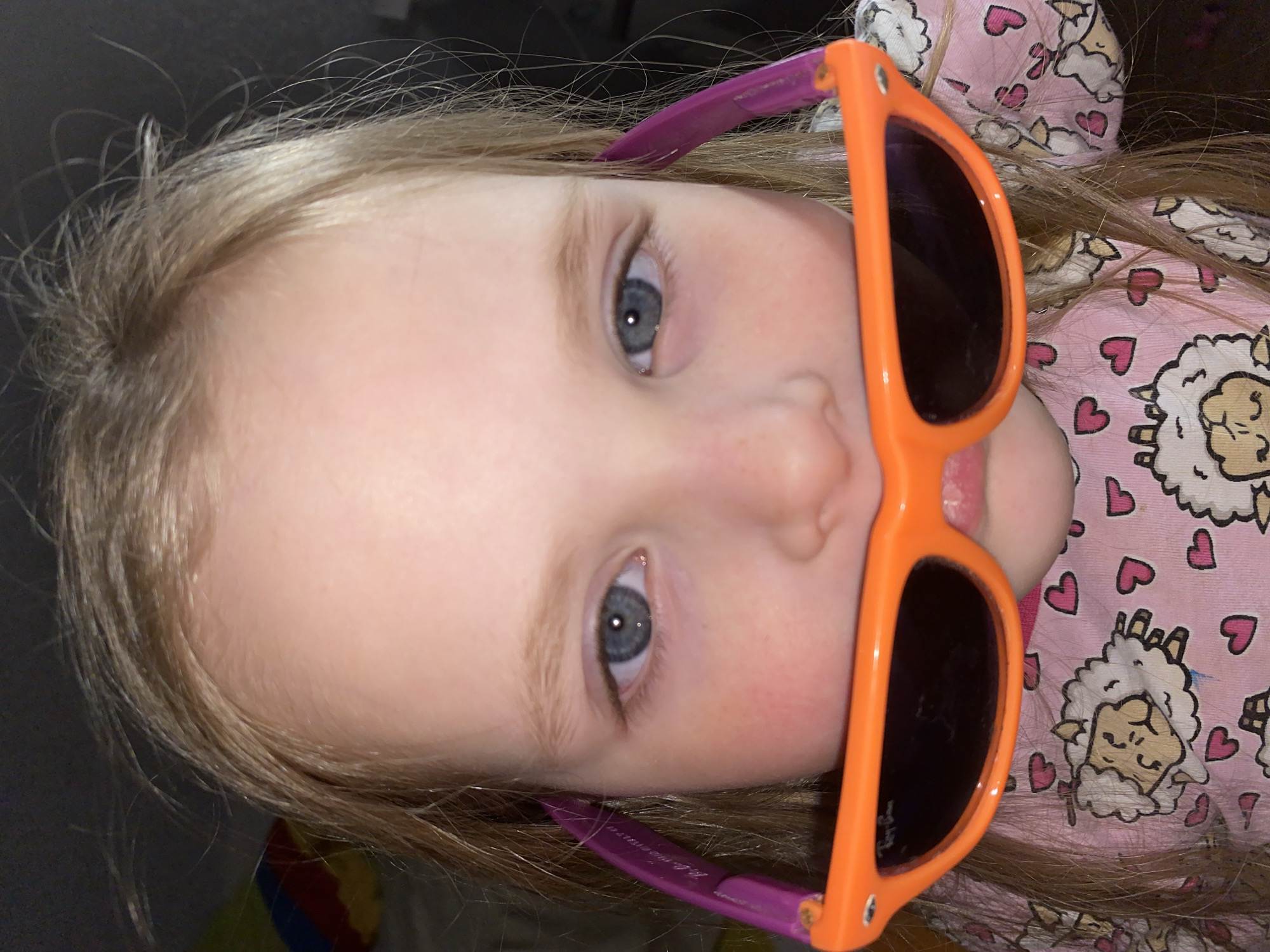 A young, light-skinned child with blonde hair and blue eyes is positioned in the center of the frame. They are wearing brightly colored orange sunglasses with purple earpieces. The child is lying down, facing upwards towards the camera. They are wearing a pink pajama top with a pattern of white sheep and pink hearts. The background is dark and out of focus. The scene is indoors and there is no evidence of this image being related to Votkinsk, Russia.