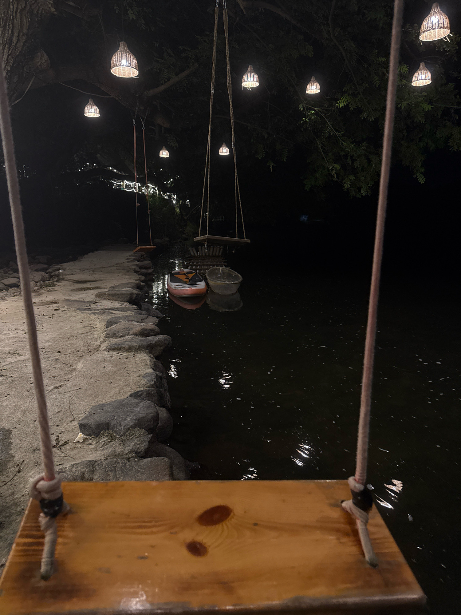 The image depicts a serene nighttime scene at a waterfront location, likely a park or recreational area, in Yiwu, China. The perspective is from a wooden swing seat, with ropes visible in the foreground, suggesting an activity area. In the background, the water is dark and reflects the ambient light. Several woven lamps hang from tree branches, casting a warm glow and illuminating the surrounding foliage. On the water, a paddleboard and a small boat are moored. A stone pathway runs alongside the water, bordered by rocks, leading towards the trees and lights. The overall atmosphere is peaceful and inviting, highlighting a tranquil evening by the water.