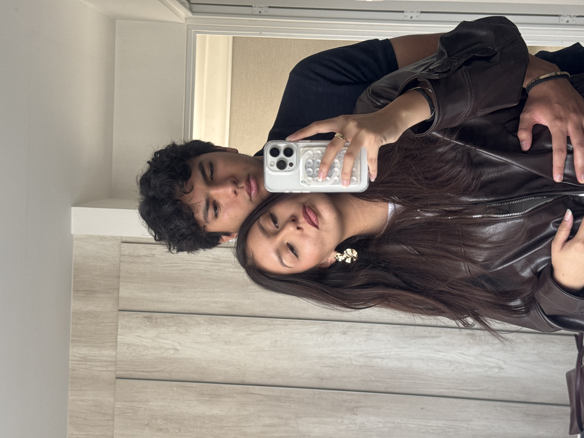 A young couple is taking a selfie in what appears to be an indoor setting, possibly a home. The man, on the left, has dark, curly hair and is wearing a black T-shirt. The woman, on the right, has long, dark hair and is wearing a brown leather jacket. She is holding a white smartphone to take the picture. A decorative gold earring is visible. The location is not discernable as being San Francisco Ocotlán (Ocotlán), Mexico from the photograph. The background includes a wall with vertical panels and a doorway leading to another room.