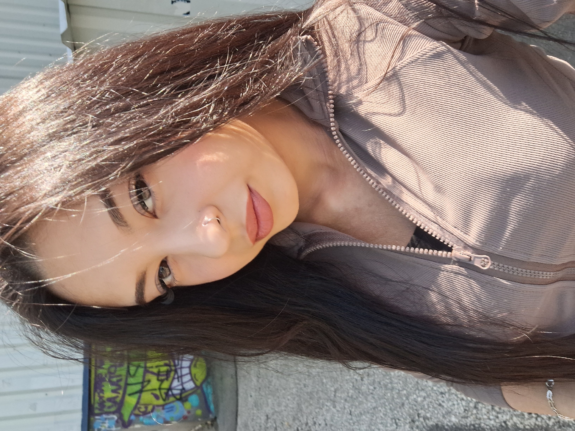 A young woman with long, dark brown hair is pictured in a close-up, angled shot. She is wearing a beige zippered jacket. The background shows a concrete surface and a partially visible graffiti-covered wall. The location appears to be an urban area within Sofia, Bulgaria.
