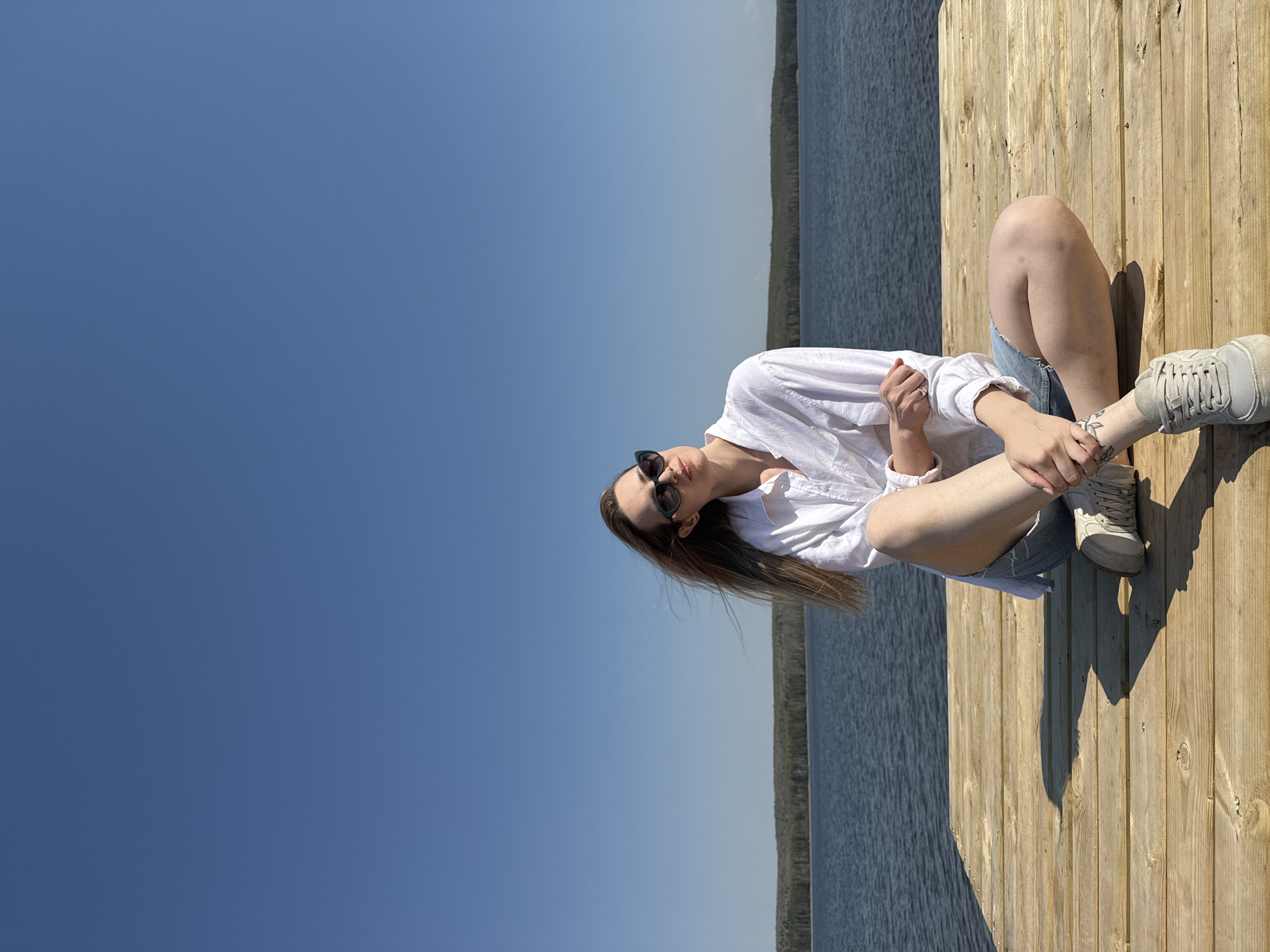A woman is sitting cross-legged on a wooden pier overlooking a body of water. She is wearing a white, button-down shirt, denim shorts, and light-colored sneakers. She has sunglasses on and her hair is down. The background includes a clear blue sky, the body of water, and a line of trees in the distance. Based on the prompt, this image was taken in Chaikovsky, Russia.