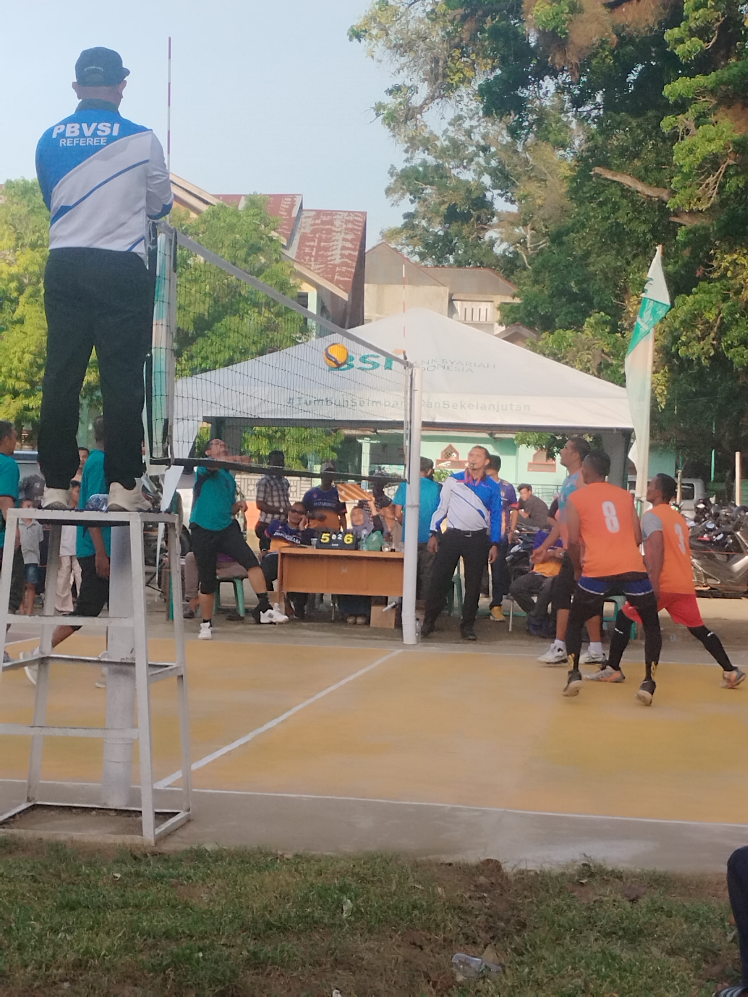 A volleyball match is underway at a court near Jalan Teuku Cik Ditiro, Lhokseumawe, Indonesia. The game is being officiated by a referee standing on a high platform, his back to the camera. He wears a blue and white uniform with "PBVSI REFEREE" printed on it. On the court, players in teal and orange uniforms are engaged in play. One player in teal is mid-air, about to hit the ball, while others in orange appear to be strategizing. Spectators and officials are visible behind a net and a large white tent, suggesting a formal event. The court surface is yellow with white lines, and the surrounding area includes trees and buildings. A score board showing "5 0 26" is also visible. The sky is clear, indicating daytime.