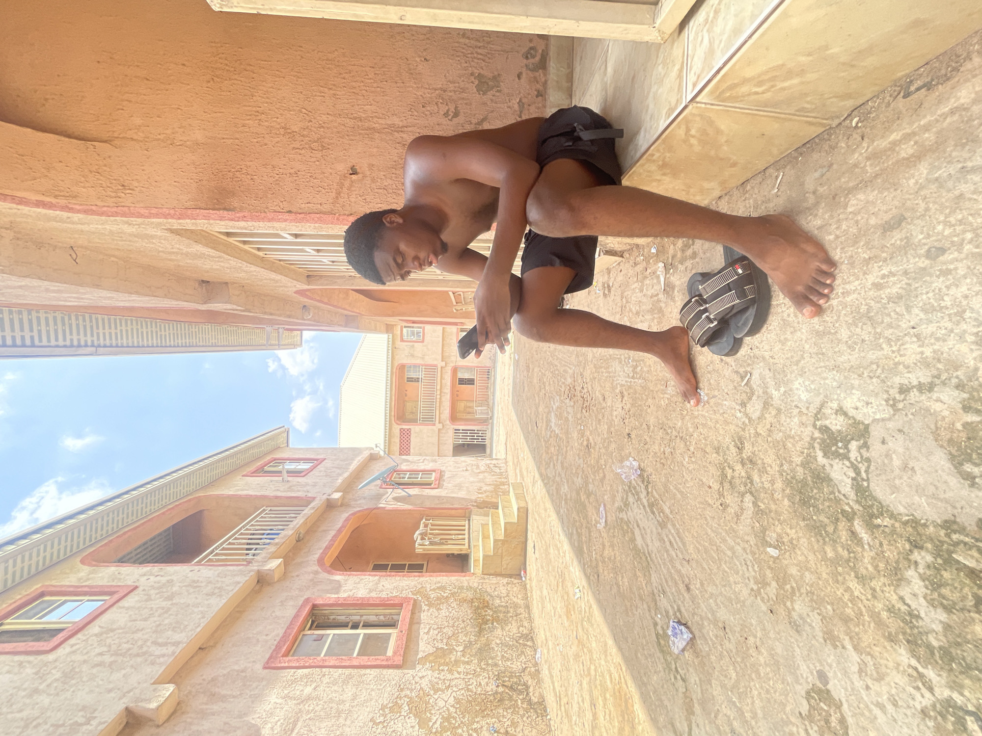 An outdoor scene captures a young man sitting on the concrete ground in an alleyway or passage between buildings, likely in Lokoja, Nigeria. The image, presented as if rotated ninety degrees, shows the man shirtless, wearing black shorts, and engrossed in his smartphone. He leans against a light-colored wall or a low step, his gaze directed downwards at the device in his hands. A pair of black sandals with white stripes rests beside his bare feet.
The surroundings consist of multi-story buildings with a warm, sandy-peach colored stucco exterior, featuring distinct windows with light-colored frames and some with red trim. Several balconies with railing details are also visible on the buildings. The concrete ground shows varied textures and some darker, possibly damp or mossy, patches, along with scattered small pieces of debris or litter. The bright blue sky, partially obscured by light clouds, indicates it is daytime, likely sunny or partly cloudy. The overall atmosphere appears casual and everyday, with the man seemingly taking a break or passing time. No discernible text is visible in the image.