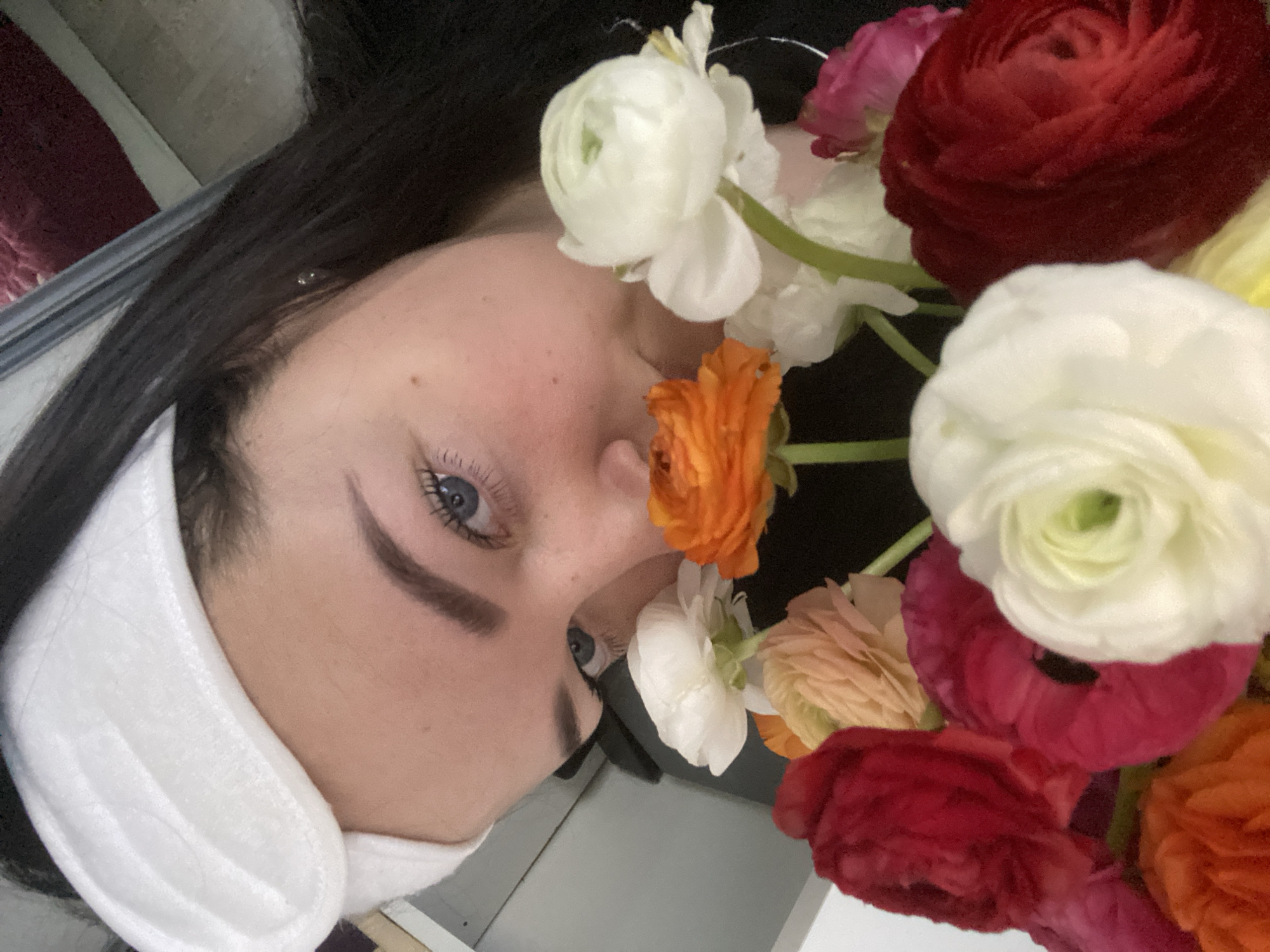 This close-up, angled portrait features a person, likely a young woman, partially framed by a vibrant bouquet of flowers. The image is oriented vertically, with the subject's head tilted. She has long, dark hair, striking light blue eyes, and well-defined eyebrows. Her fair skin shows a few small moles on her forehead. A white, padded accessory, possibly a mask or earmuffs, covers the lower part of her face and ear.
A profusion of colorful Ranunculus flowers surrounds her face, particularly on the right side of the frame. The bouquet includes dense blooms in various hues: prominent white flowers, a bright orange bloom near her nose, and deep red, fuchsia pink, and pale peach/yellow flowers intermingled throughout. The flowers appear fresh and are positioned very close to her.
The activity depicted is a close-up portrait or self-portrait, emphasizing the subject's face amidst the natural beauty of the blossoms. Her visible eye conveys a calm and serene expression. The lighting is soft, suggesting an indoor setting, and the background is indistinct and dark, drawing all focus to the subject and the floral arrangement. This scene is set in Sochi, Russia. No discernible text is visible in the image.
