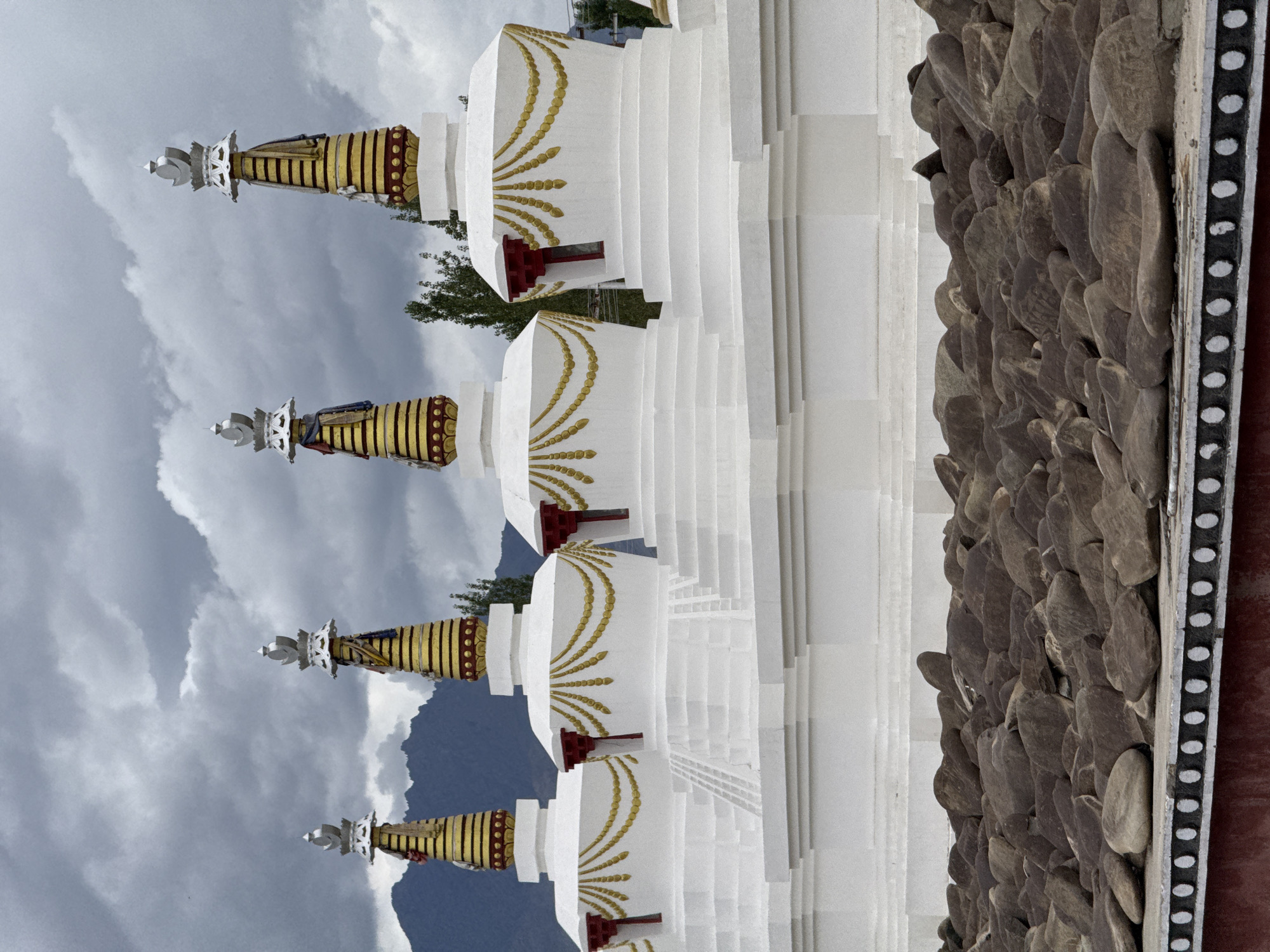 The image shows a row of white stupas in Thiksey, India, under a cloudy sky. The stupas are tiered, with each level resembling steps. The upper portions of the stupas are decorated with gold and red accents, and prayer flags hang from some of them. In the foreground, a collection of brown, irregularly shaped rocks fills the space in front of the stupas. Behind the stupas, a dark mountain range is visible, and several trees are scattered throughout the landscape. The overall scene is serene and appears to be a religious site.