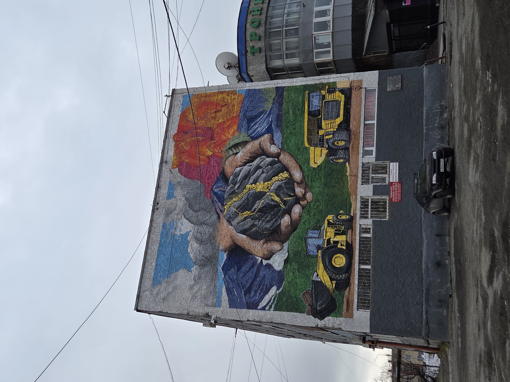 A vertical shot of a mural on the side of a building in Unknown, Unknown. The mural depicts two hands holding a piece of what appears to be coal or ore. There are two bulldozers painted on the right side of the mural. The hands holding the coal/ore are painted with bright colors in the background. A car is parked next to the building.