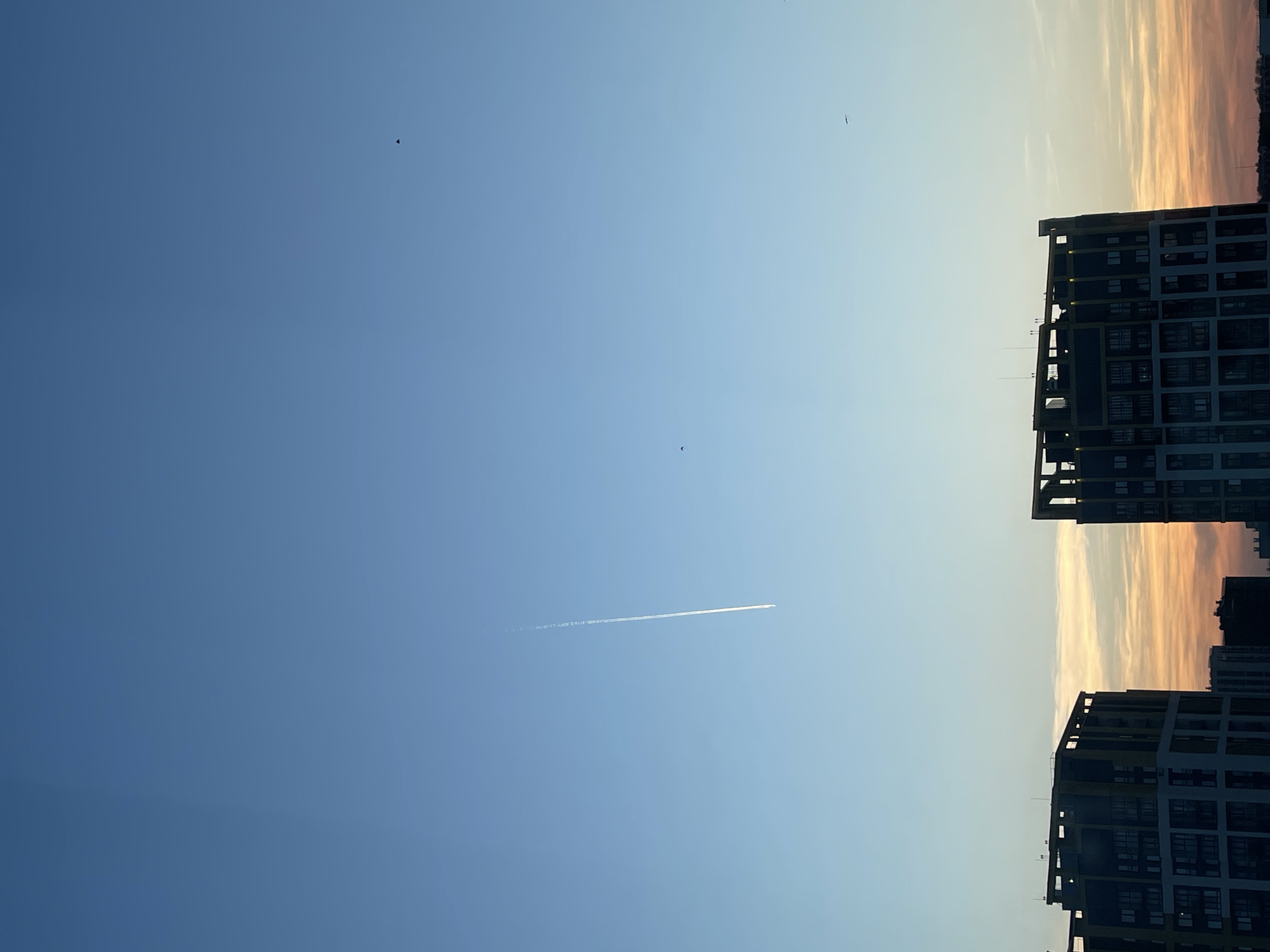 The scene presents an urban setting in Izhevsk, Russia, under either sunrise or sunset lighting conditions. The upper sky displays a gradient from deep blue overhead to pale blue, transitioning to orange and yellow at the horizon. A singular, white aircraft contrail extends vertically downward from the upper-middle left. Multiple small, dark aerial objects, consistent with birds or distant aircraft, are visible against the sky; one triangular object is noted in the upper-right. The lower frame is occupied by the dark silhouettes of several modern, multi-story buildings. Two primary structures, one on the left and one on the right, feature multi-windowed facades and prominent skeletal upper sections. A lower-lying urban profile is visible between these buildings, silhouetted against the horizon. No individuals are discernible.