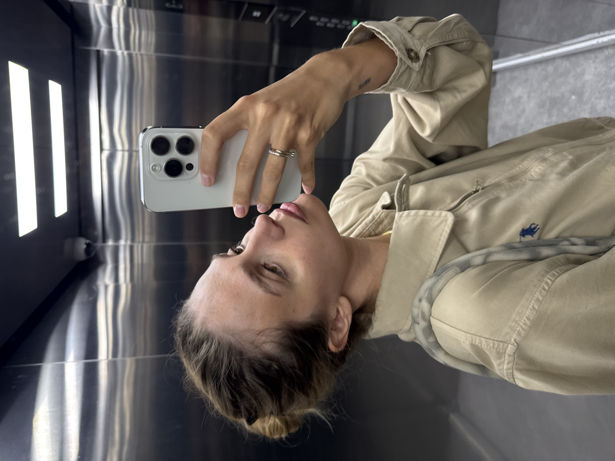 A person is taking a selfie with a white smartphone in an elevator. The person is wearing a light brown jacket. The elevator walls are made of stainless steel. The location is unknown.