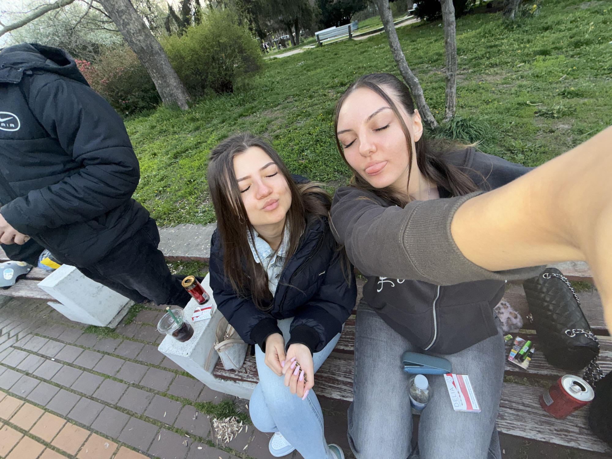Two young women are sitting on a concrete bench in a park, posing for a selfie. One woman on the right sticks her tongue out. There is a man on the left of the women. The environment is a park with grass, trees, and a sidewalk. On the bench, beside the women, are cigarettes, a drink, and a can. The location may be in Sofia, Bulgaria.