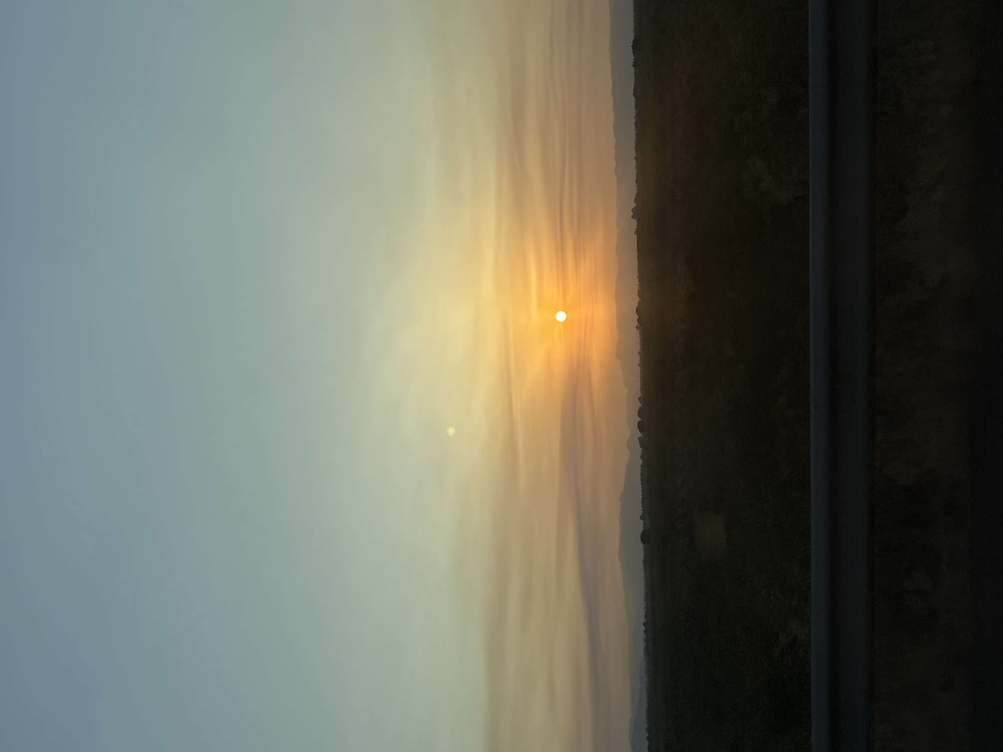 The image depicts a sunset over a rural landscape, likely viewed from a roadway. The sky transitions from a pale blue at the top to a warm orange and yellow near the horizon, with wispy clouds catching the light. The sun is a bright, small orb positioned slightly to the left of the center, casting a warm glow. In the mid-ground, a dark, flat expanse of land stretches across the frame, dotted with vegetation or low-lying trees. Distant mountains form a hazy silhouette against the sky. In the foreground, a dark, metallic guardrail runs horizontally across the bottom of the image, suggesting the perspective of someone observing the scene from a vehicle. The overall impression is one of natural beauty and tranquility. The image does not contain any information that allows for identification of its location in Vinaròs, Spain.