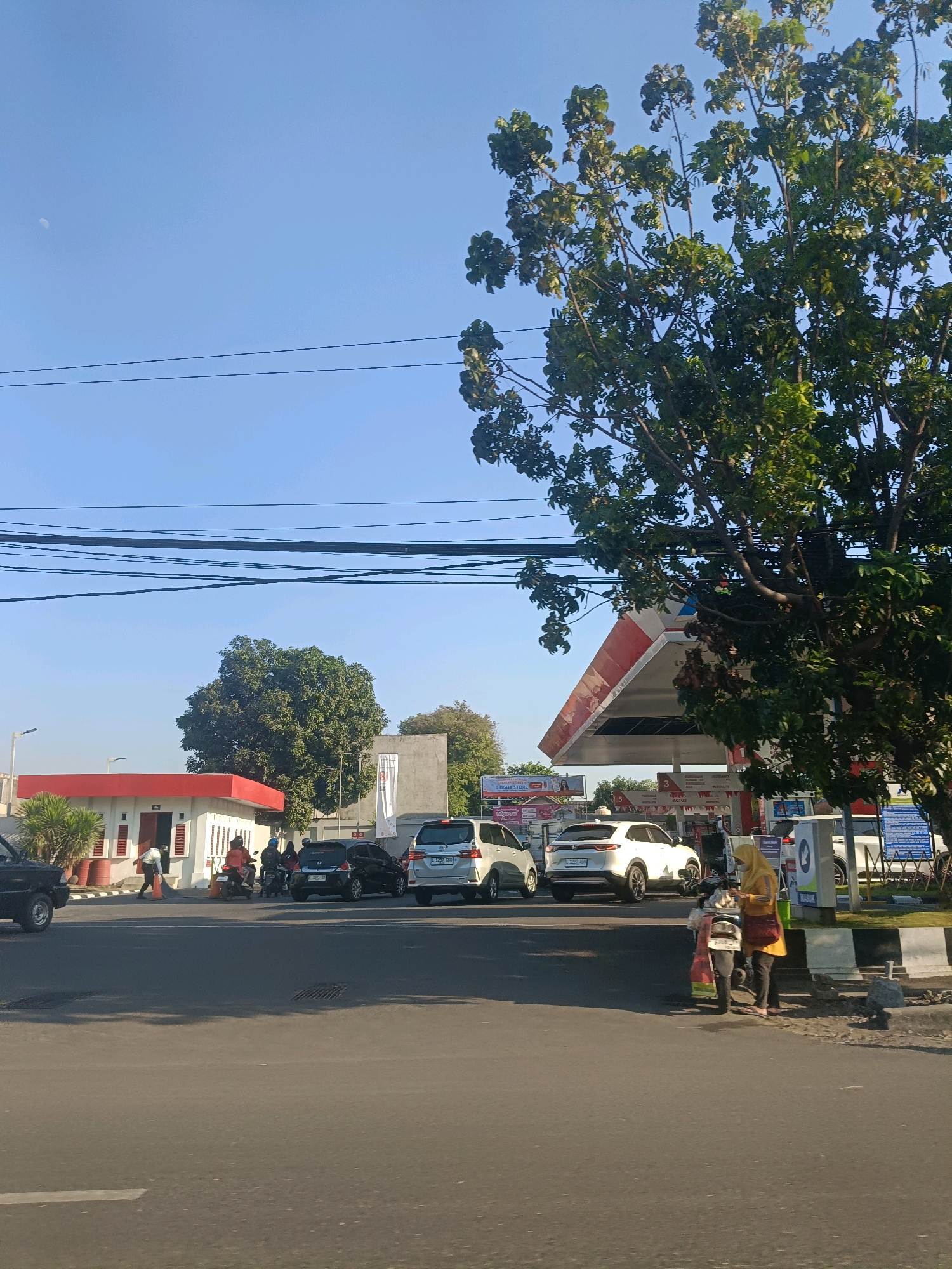 This is a sunny, daytime, eye-level, outdoor shot of a gas station in the city of Surabaya, Indonesia. The gas station, which appears to be a Pertamina station, features a red and white canopy over the pumps. Several cars and motorcycles are present, some at the pumps, and others waiting in line. One woman in a yellow hijab stands next to a motorcycle, seemingly waiting. The sky is clear and blue with the moon visible. In the foreground, the street and sidewalk are visible, with a large tree on the right side of the frame partially obscuring the gas station canopy. There are power lines running across the top of the image.