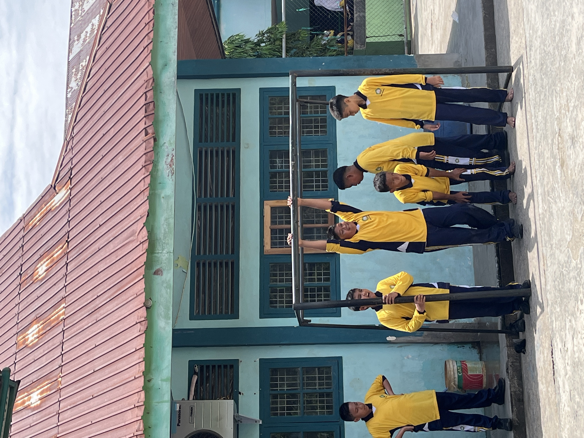 The image captures a group of boys in yellow and navy blue school uniforms standing outside a building with a turquoise-painted wall and a corrugated red roof. The building has dark blue-framed windows with intricate grates and louvered vents. A prominent metal bar structure, likely a pull-up bar, is positioned in the foreground, with one boy actively hanging from it, his arms extended upwards. Other boys are gathered around the structure, some looking at the boy on the bar, one holding onto a vertical support pole, and others standing and observing. A white Panasonic air conditioning unit is mounted on the wall to the left. On the ground, near the boys, are several dark objects that appear to be shoes and a black mat. To the left of the structure, a cylindrical container with red and white markings is visible. The ground surface is a light-colored paved area, and a green chain-link fence can be seen in the background. The scene appears to be an outdoor schoolyard or recreational area, likely in Lhokseumawe, Indonesia, given the context. The sky above is partly cloudy.