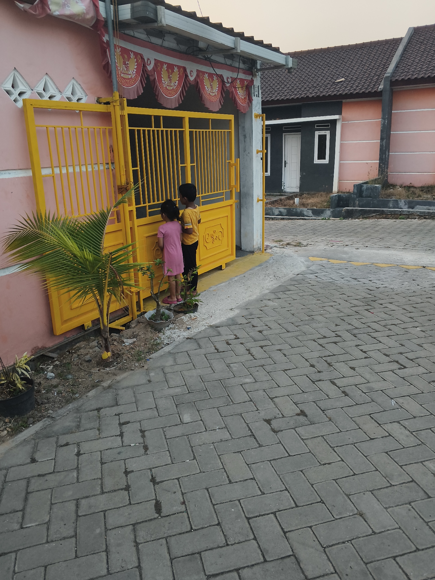 Two young children, a boy in a yellow t-shirt and a girl in a pink dress, stand facing a bright yellow metal gate on the grounds of a residential complex. The boy is on the right, and the girl is to his left. They appear to be looking through the gate into a yard. The gate is part of a pink wall. In front of the gate, to the left, is a small palm tree in a planter, and to the right, there are two smaller plants in pots. The ground in front of the gate is covered with grey interlocking bricks. Behind the gate, a street or pathway also paved with interlocking bricks extends away from the viewer. In the background, several houses with dark tiled roofs and light-colored walls are visible, along with some sparse vegetation. The sky is overcast. The scene appears to be a residential area in Banjar Agung, Indonesia.
