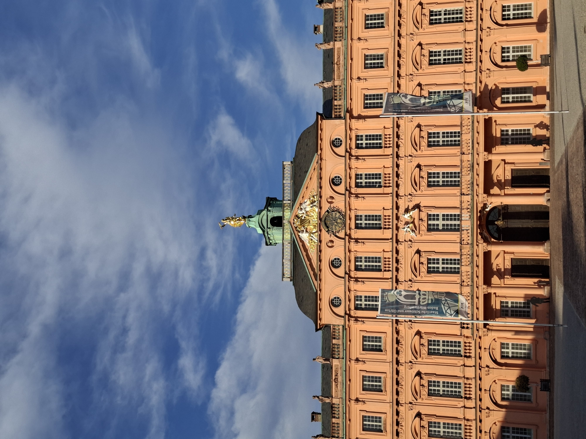 This is a low-angle, full-frame shot of the Rastatt Residential Palace in Rastatt, Germany. The palace, painted in a warm peach hue, dominates the lower two-thirds of the frame. Its facade features a symmetrical design with multiple rows of windows, ornate architectural details, and a central entrance. Above the central section, a prominent clock face is visible. At the very top, a golden statue stands atop a green, domed structure, set against a bright blue sky with scattered white clouds. Two vertical banners hang in front of the palace, displaying text and graphics. In the foreground, paving stones form a courtyard. A lone figure stands near the entrance, casting a shadow. Small, sculpted trees are placed on either side of the central entrance, and another is visible in a planter to the left. The image captures a sunny day, with shadows indicating the time of day.