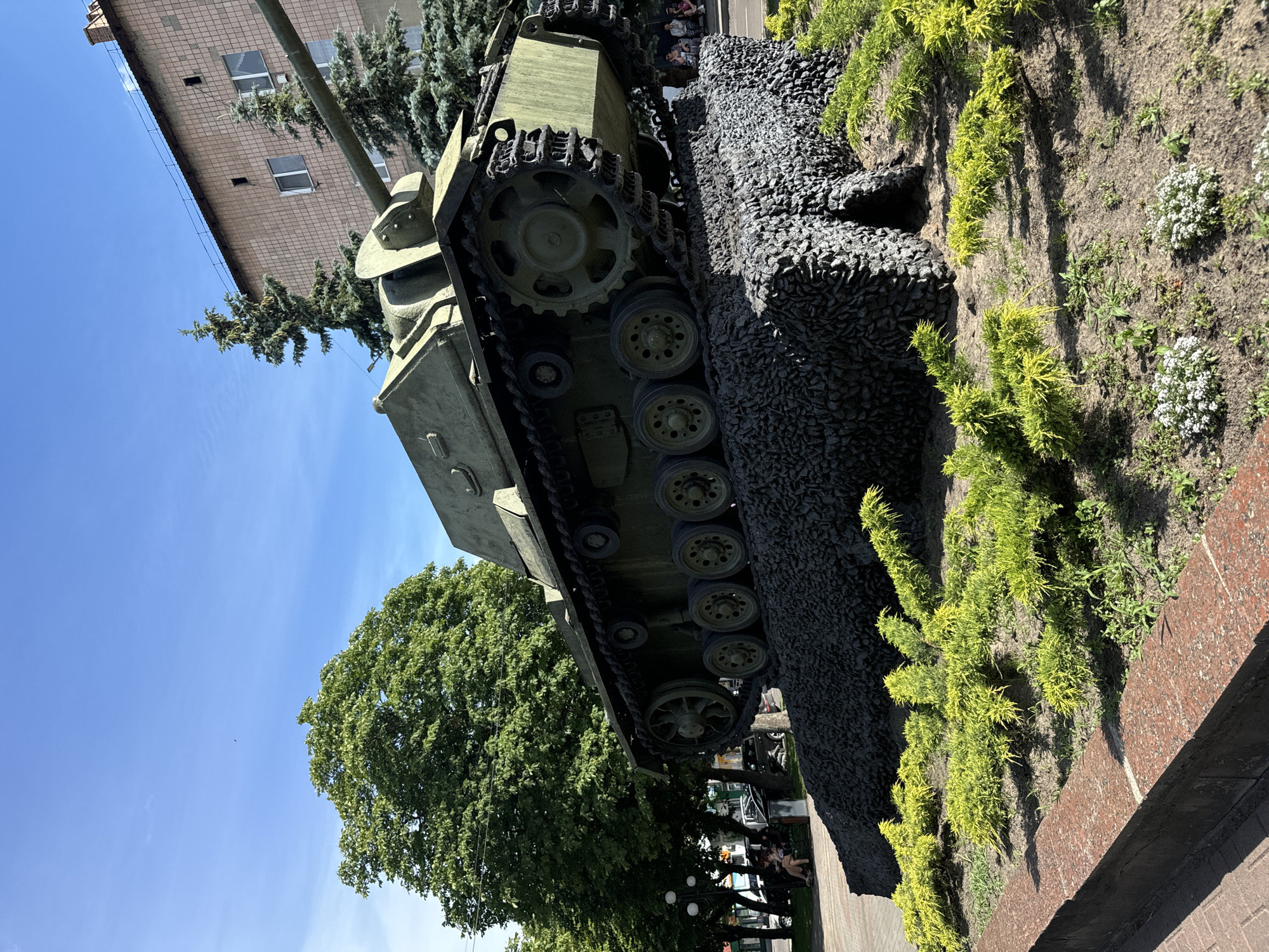 The image shows a close-up, low-angle view of a military tank, oriented vertically. The tank is green and appears to be a monument, as it is situated in a landscaped area with dark grey rocks and green plants. The tank's tracks and wheels are clearly visible. Behind the tank, a building with a tiled facade and several windows can be seen. A large green tree with dense foliage is also present, and a clear blue sky with a few wispy clouds occupies a significant portion of the frame. The lighting suggests it is daytime, likely late morning or afternoon, with bright sunlight casting shadows.
In the background, beyond the main subject, there are glimpses of people walking and sitting, suggesting a public space or park. The overall scene conveys a sense of outdoor ambiance, possibly a memorial site or a display of historical military equipment in Sarny, Ukraine. No visible text is discernible in the image.