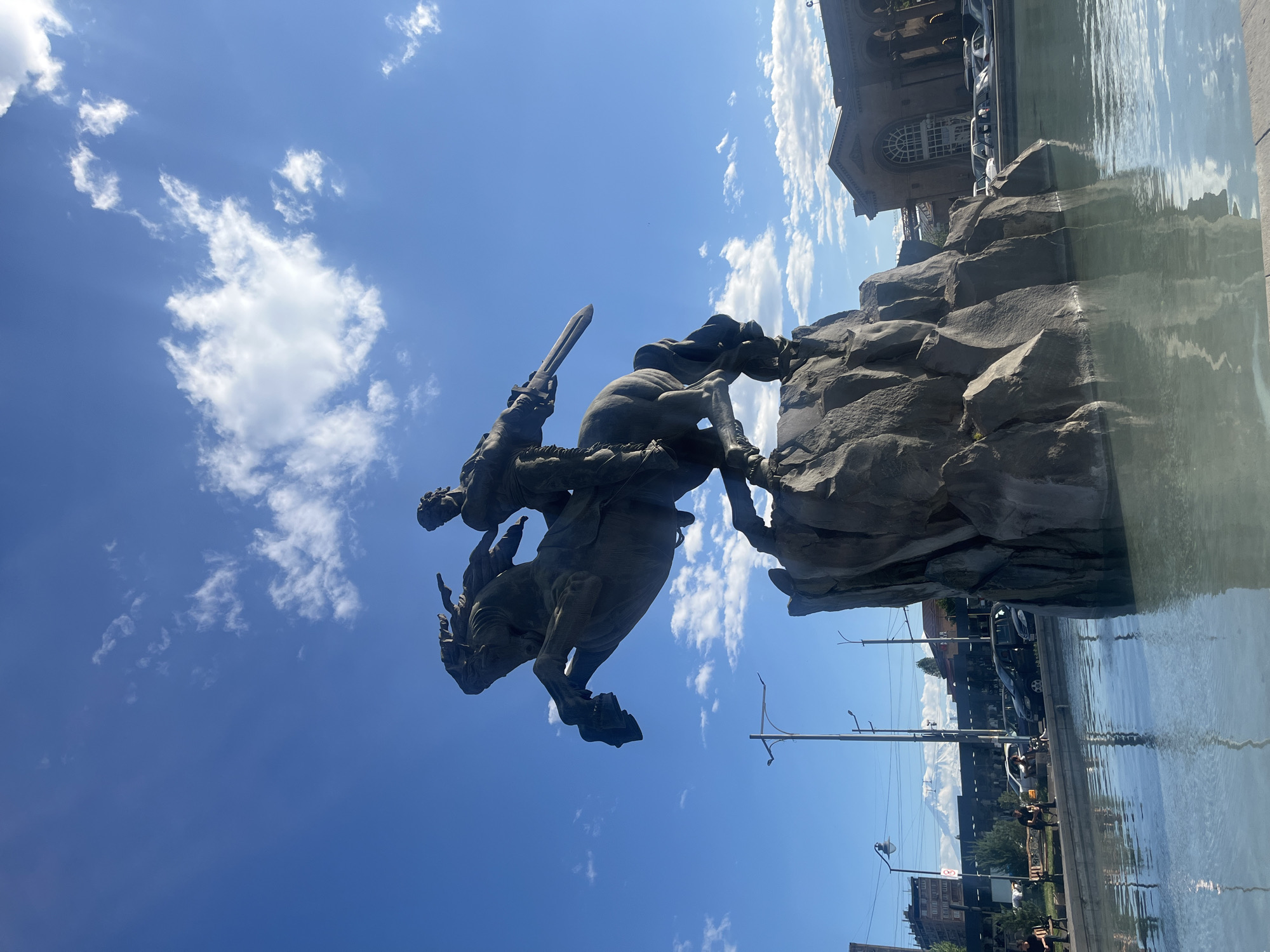 The image shows the statue of Sasuntsi Davit in Yerevan, Armenia. The statue is bronze and depicts Davit on horseback, raising a sword. The statue is situated atop a rocky pedestal with a fountain surrounding it. The backdrop is a blue sky with scattered clouds. In the background, there are buildings, cars, and other urban elements. The statue is located at the center of Yerevan's main square. The shot appears to have been taken from a low angle, possibly at the edge of the fountain.