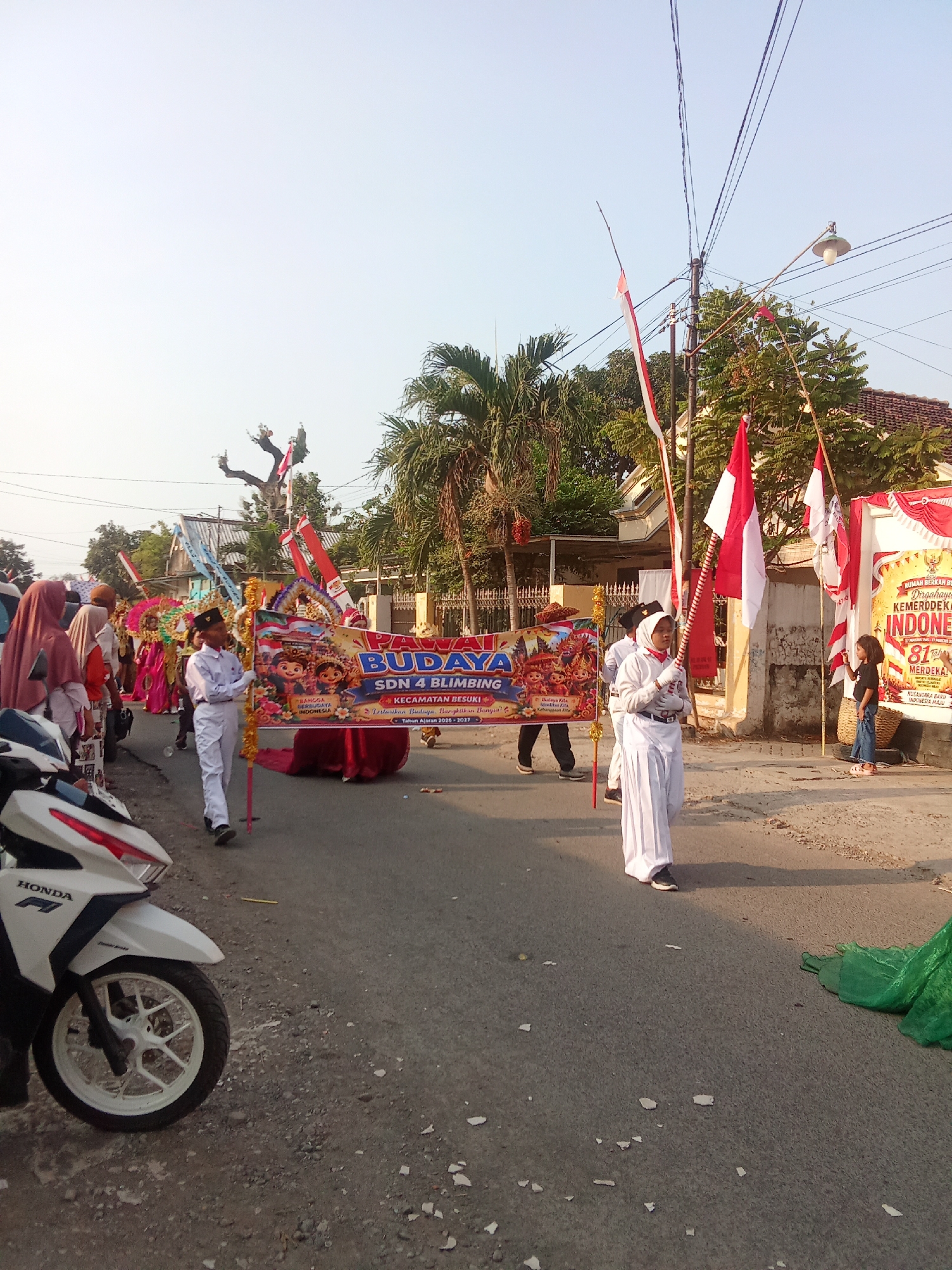 A group of children in white uniforms are participating in a parade on a street. The children are holding a banner that reads "BUDAYA SDN 4 BLIMBING" and "KECAMATAN BESUKI". They are also holding Indonesian flags. There are other people in the background, some dressed in traditional clothing. A white motorcycle is parked on the left side of the image. The street is paved and there are trees and houses on either side. The sky is clear and blue. The event appears to be a cultural or national celebration.