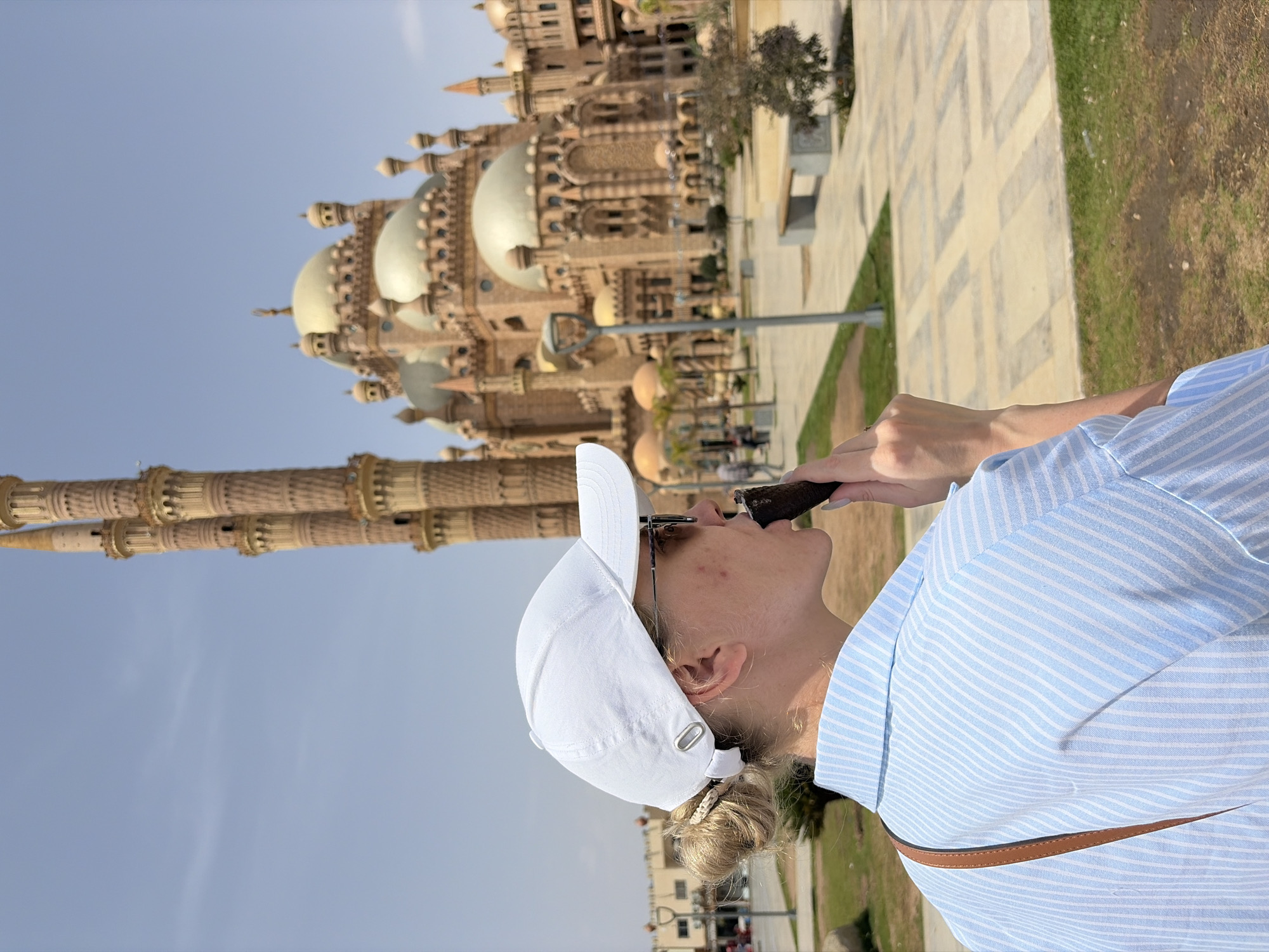 The image captures a woman in Sharm El Sheikh, Egypt, with the Al-Mustafa Mosque as a backdrop. The woman, wearing a white cap and a light blue striped shirt, is holding a dark-colored ice cream cone to her mouth. She appears to be enjoying the treat with her head tilted back. The mosque, an ornate structure with multiple domes and minarets, occupies a significant portion of the background, showcasing its architectural details. A manicured lawn and paved walkways are visible on the right, indicating a well-maintained outdoor space. The sky is a clear blue, suggesting a sunny day.
