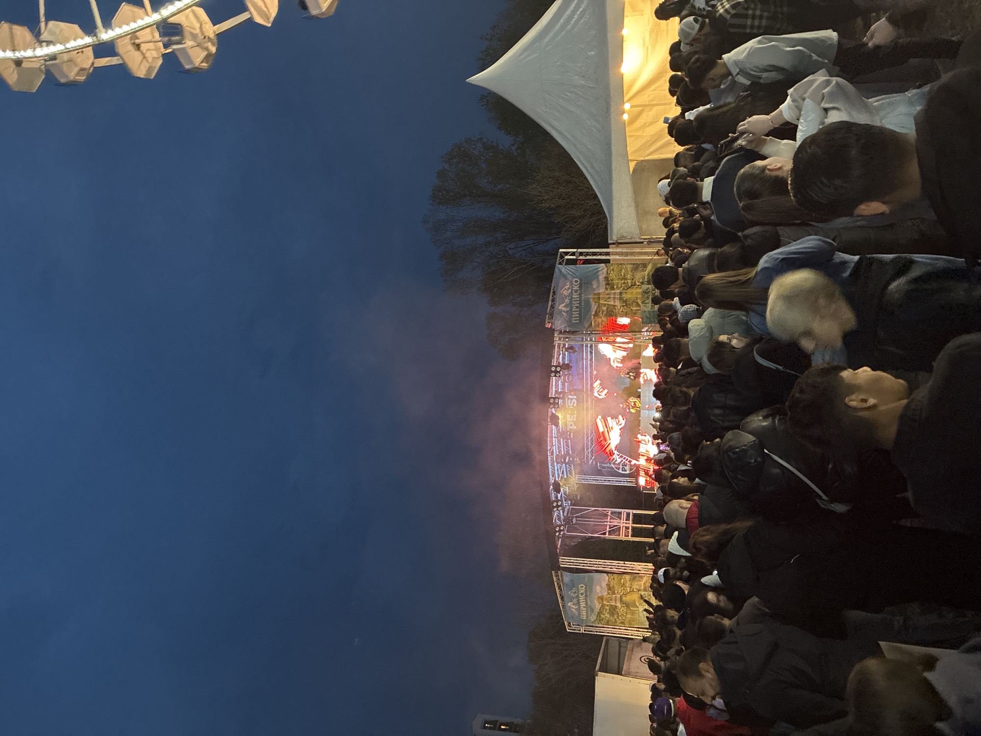 A sideways, upward-angled shot depicts a crowd of people gathered for an outdoor event in Plovdiv, Bulgaria. A stage is visible, illuminated with red and white lights, surrounded by a structure with signage in Cyrillic script; one sign appears to say "ПИРИНСКО" (Pirin in Cyrillic). Above the stage, trees and a large white tent are visible. In the background, a Ferris wheel's structure is visible against a dark blue sky. The focus is on the concertgoers, mostly dark-haired individuals of varying heights, angled as though facing the stage. Clothing ranges from dark jackets to lighter-colored tops.