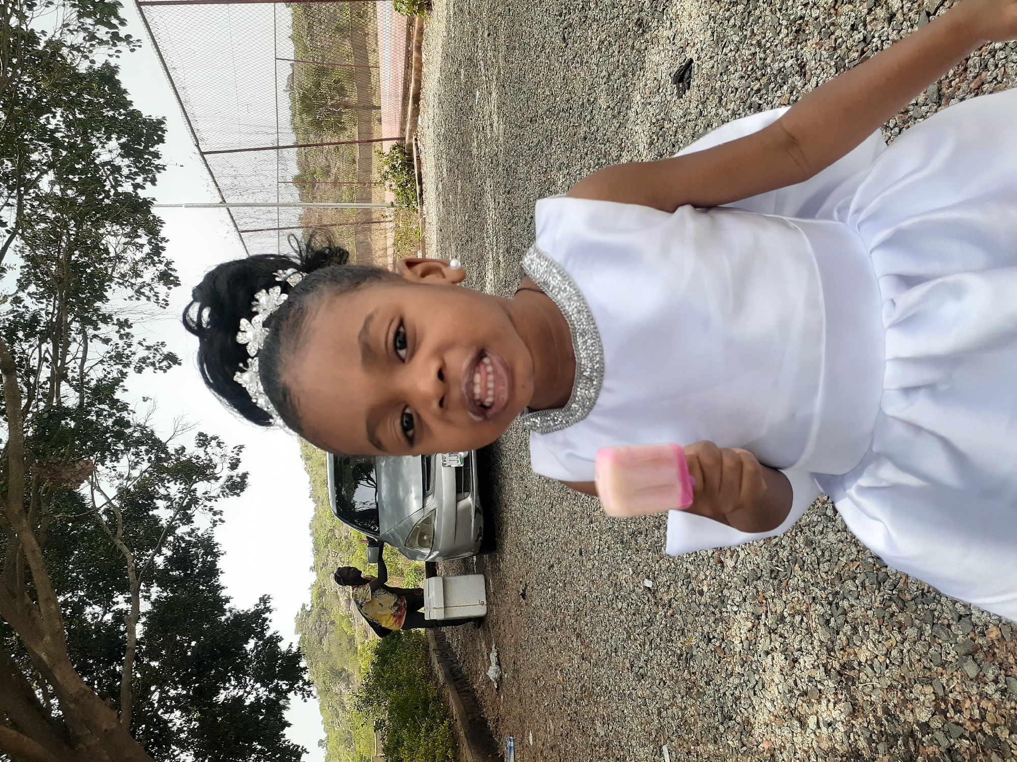 A child in a white dress is holding a pink popsicle. The child is standing on a gravel surface, with a car and a person standing next to a cooler in the background. A tree is on the left and a fence is in the background. The location is the city of Unknown, Unknown.