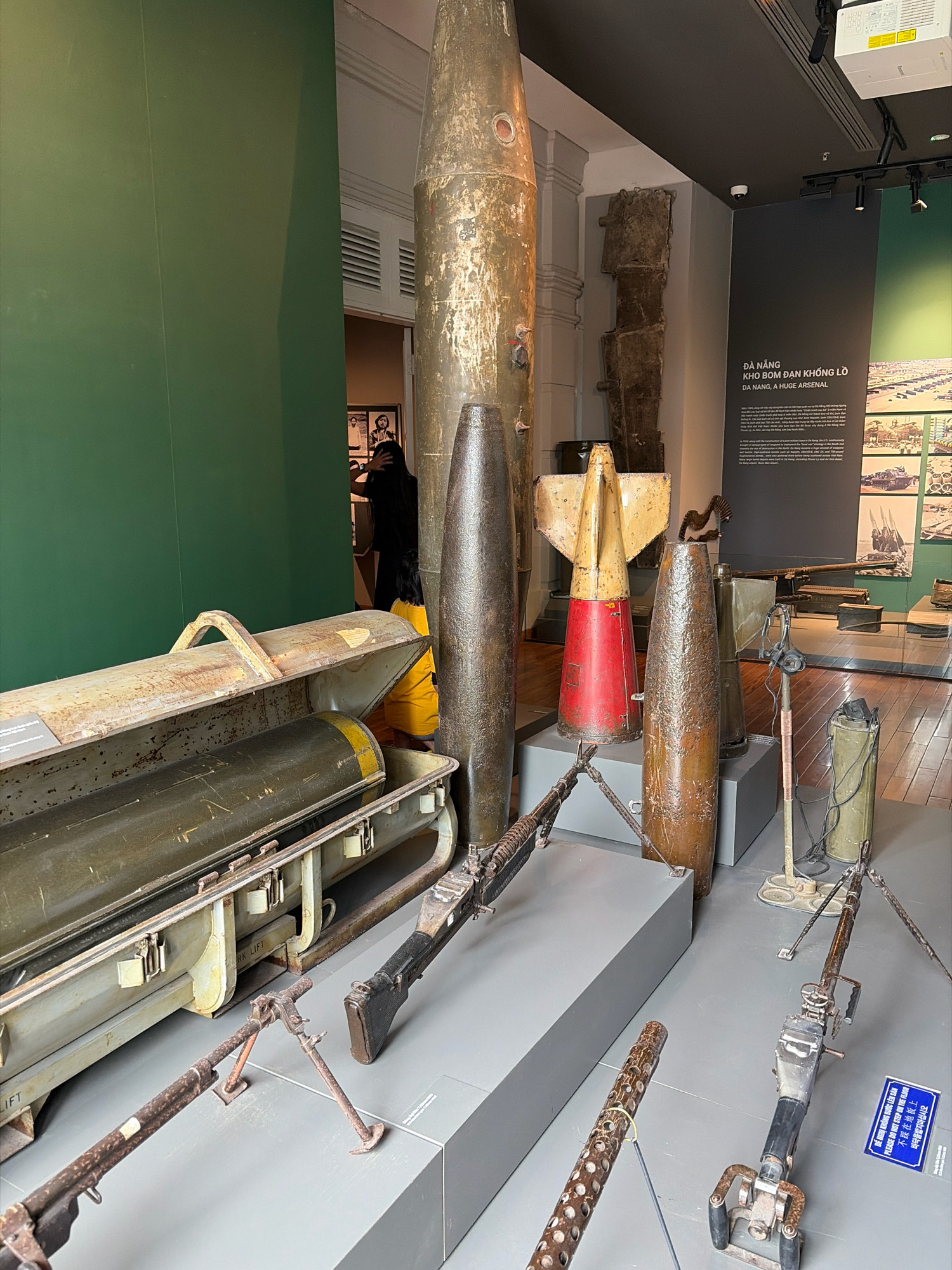 The image depicts an exhibition at the Da Nang Museum, Vietnam, showcasing a collection of military artifacts related to the Vietnam War. In the foreground, a large, cylindrical bomb casing rests horizontally within a weathered, open container. Adjacent to it, another large bomb casing stands vertically. To the right of these, a mortar round, painted red and yellow, is positioned next to a tall, rusty bomb casing. Further to the right, a heavy machine gun with a bipod is displayed on a pedestal, with a separate machine gun barrel visible in the lower left corner. Behind these artifacts, a large, dark green wall serves as a backdrop, with a display panel on the right featuring Vietnamese and English text, and an accompanying photograph of military equipment. In the background, a section of the museum's interior is visible, including a dimly lit doorway where a person, dressed in black, appears to be looking at exhibits. The overall environment suggests a museum exhibit focused on wartime history, with a somber and informative presentation of historical military hardware.