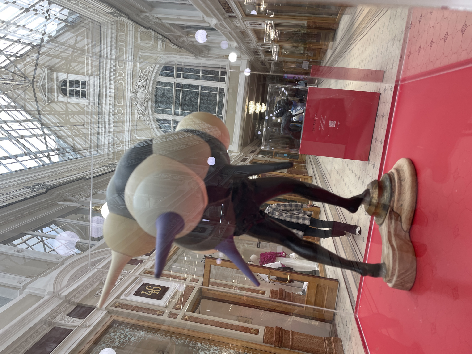 This image shows a statue in the Singer House (Dom Knigi) bookstore in Saint Petersburg, Russia. The statue is a sculpture of a figure with a dark, shiny body and a large, balloon-like head composed of tan and gray segments with purple tips. The figure is standing on a wooden base. The statue is displayed behind glass, and the reflection of the store's interior, including an ornate arched ceiling with glass panels and a chandelier, is visible. In the background, there are display windows with mannequins, shoppers, and more reflections. The floor of the store is tiled in a geometric pattern, and the statue is placed on a red platform.