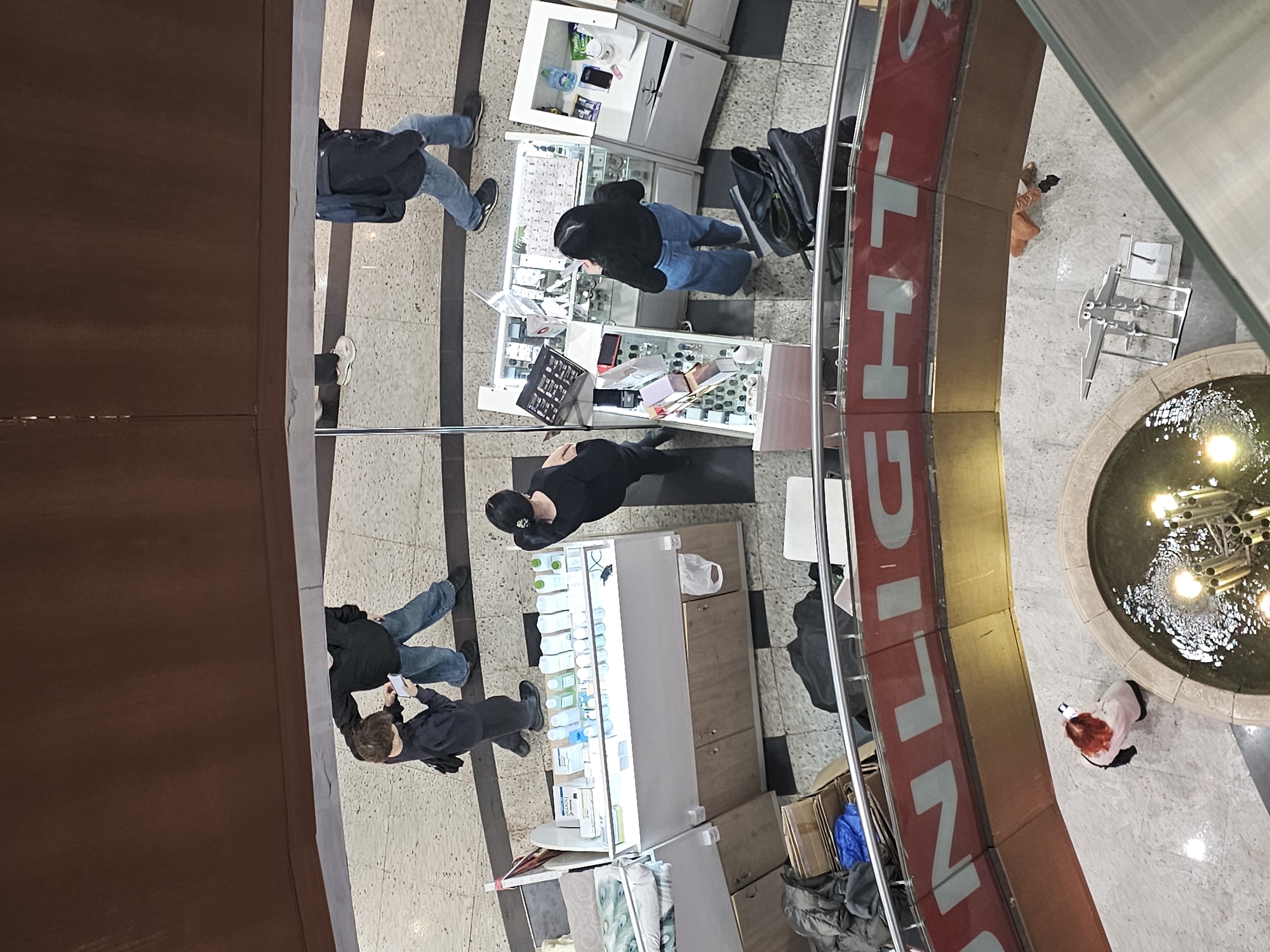 The image, taken from an elevated perspective, shows the interior of a shopping mall. The scene is in the city of Khimki, Russia. Several retail kiosks are visible, displaying items like cosmetics, jewelry, and accessories. People are standing at these kiosks, seemingly browsing or shopping. Some individuals appear to be in casual attire, with one notable figure wearing a bright red wig and a white coat near a fountain. A fountain with illuminated water features is positioned near a curved balcony. The mall floor is tiled and marked with dark lines. The edge of the balcony is lined with red lettering. Boxes and bags are stacked near the edge of the frame. The image captures the hustle and bustle of shoppers within a commercial environment.