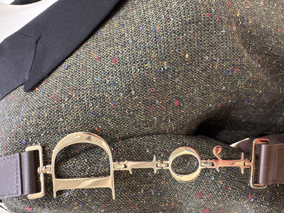 The image displays a close-up view of a luxury fashion item and its accessory, consistent with a retail display or showroom environment in Paris, France. A dark olive-green tweed garment, likely a jacket or blazer, occupies the majority of the frame. The fabric features a tight weave with numerous interwoven, multi-colored flecks in shades of red, blue, yellow, purple, and white, contributing to a speckled texture. A sharp, black lapel is visible on the upper right side of the garment. Overlaying the garment is a prominent gold-toned metallic accessory. This accessory forms the brand name "DIOR" using individually cast and linked letters: "D," "I," "O," and a lowercase "r." The "D" is connected to a dark brown leather strap at the top, and the "r" is connected to another dark brown leather strap at the bottom, indicating it functions as part of a bag strap or a decorative charm. No individuals are visible in the scene.