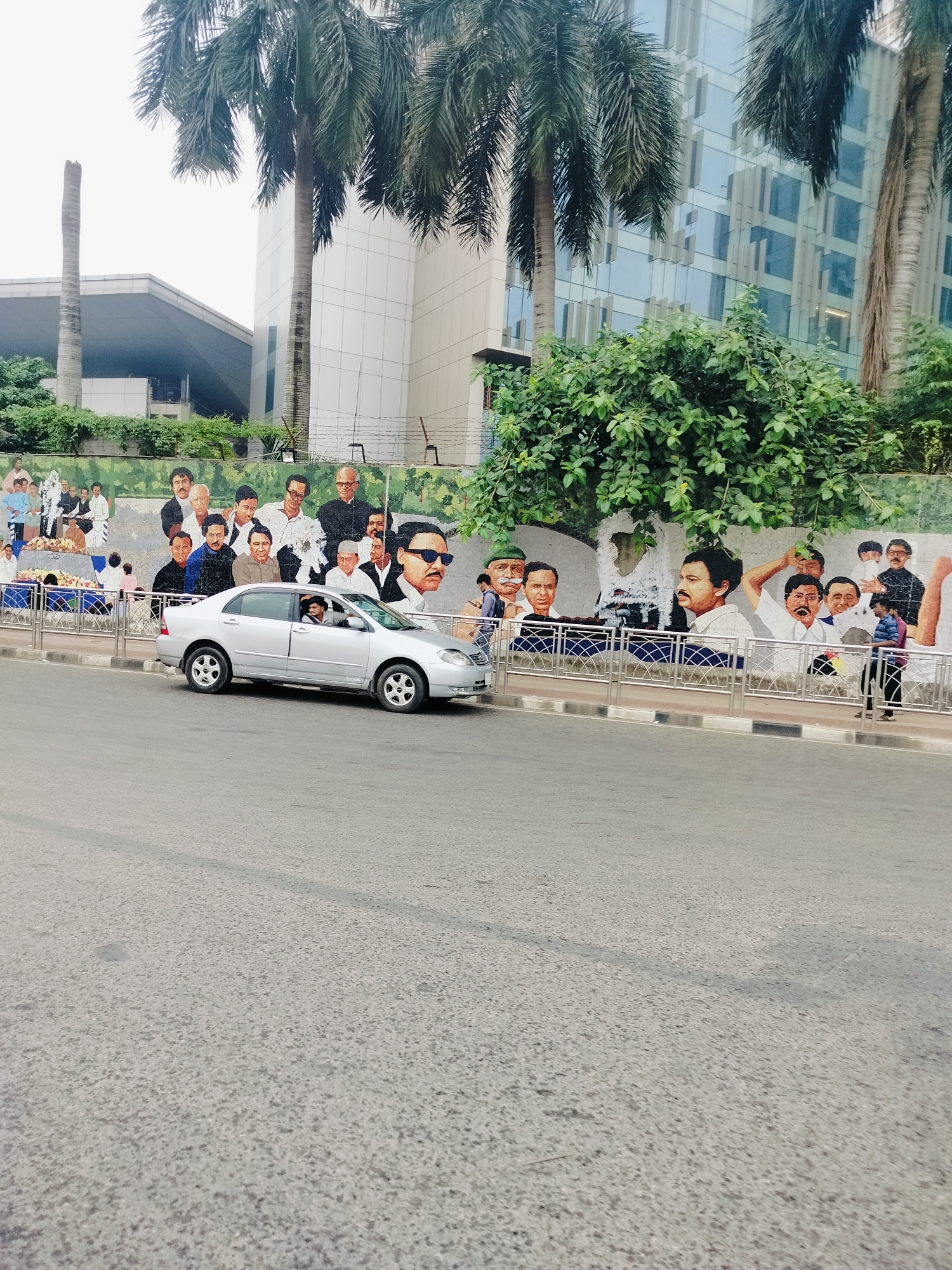 A silver sedan is parked on the side of a road, with a driver visible inside. Behind the car, a mural adorns a wall, depicting a collection of portraits of various men, some in traditional attire and others in suits. The mural is partially obscured by lush green foliage and tall palm trees. In the background, a modern glass-fronted building rises, reflecting the sky. The overall scene appears to be a public space in Dhaka, Bangladesh, with the mural likely representing historical or notable figures.