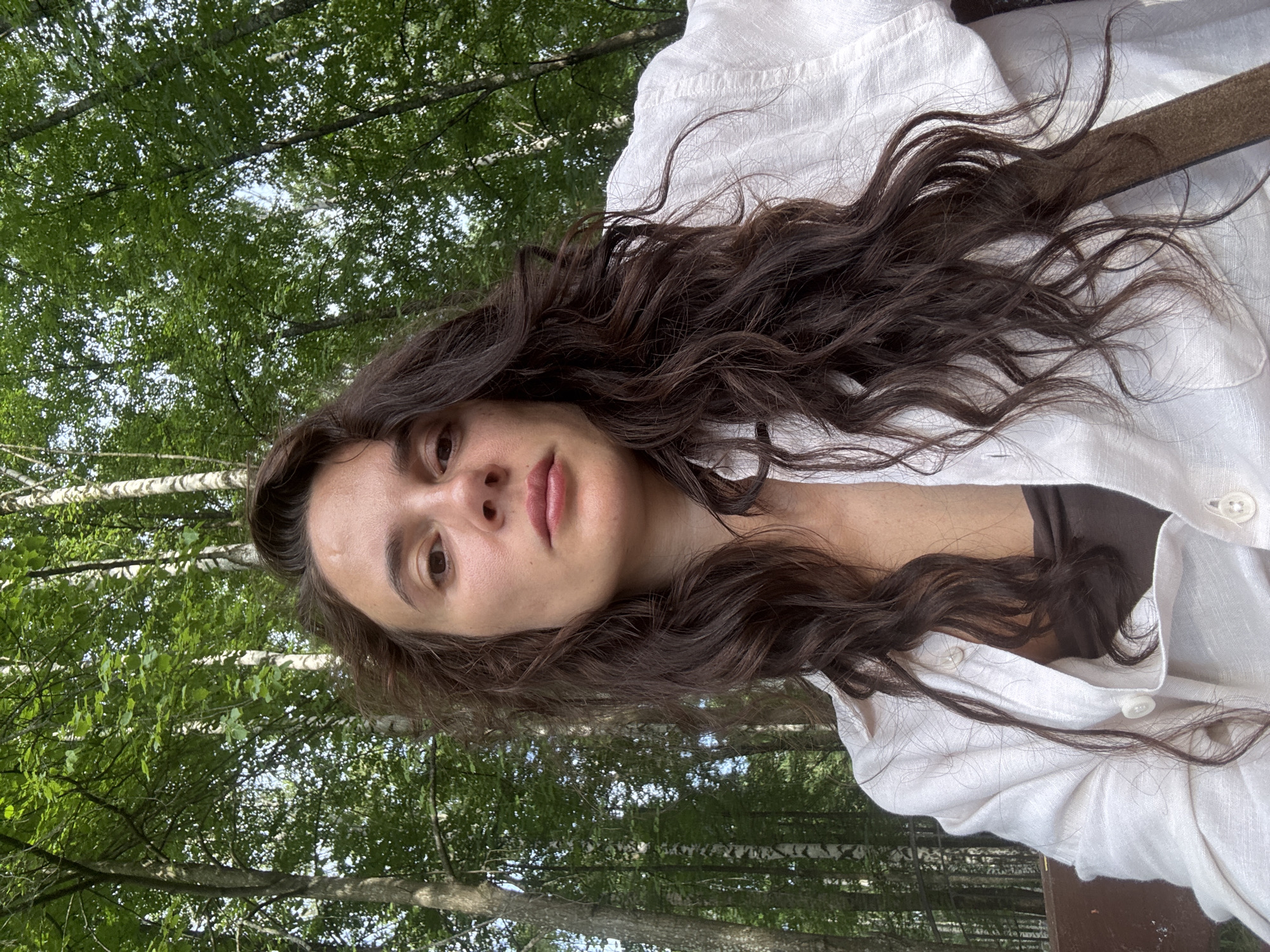 A medium shot captures a woman with long, dark, wavy hair from a low angle, looking directly at the camera. She is wearing a loose, unbuttoned white linen shirt over a dark brown top. Her face is framed by her hair, which falls over her shoulders. The background consists of a dense forest with tall trees, some appearing to be birch trees with white bark, and lush green foliage. The lighting suggests an outdoor setting during the day, with sunlight filtering through the leaves. The image appears to have been taken in the city of Chaikovsky, Russia, given the presence of birch trees common in the region and the overall natural environment.