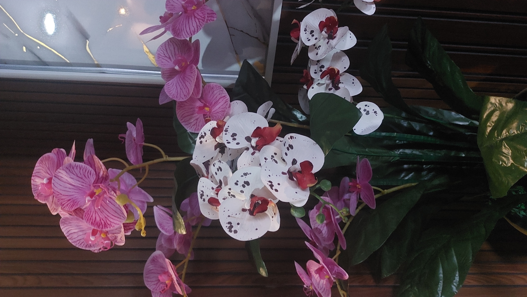 The image shows a close-up of a floral arrangement featuring several artificial orchids. The arrangement includes clusters of pink orchids with darker pink stripes, and white orchids with dark red centers and speckled petals. Large, dark green artificial leaves are also prominent, forming the base of the arrangement. The background consists of vertical wooden panels and a portion of a framed artwork with white and gold abstract patterns. The lighting suggests an indoor setting, likely a decorative piece in a residential or commercial space. No people are visible, and there are no interactions to describe. The specific location within the City of Medan, Indonesia, cannot be determined from the image alone.