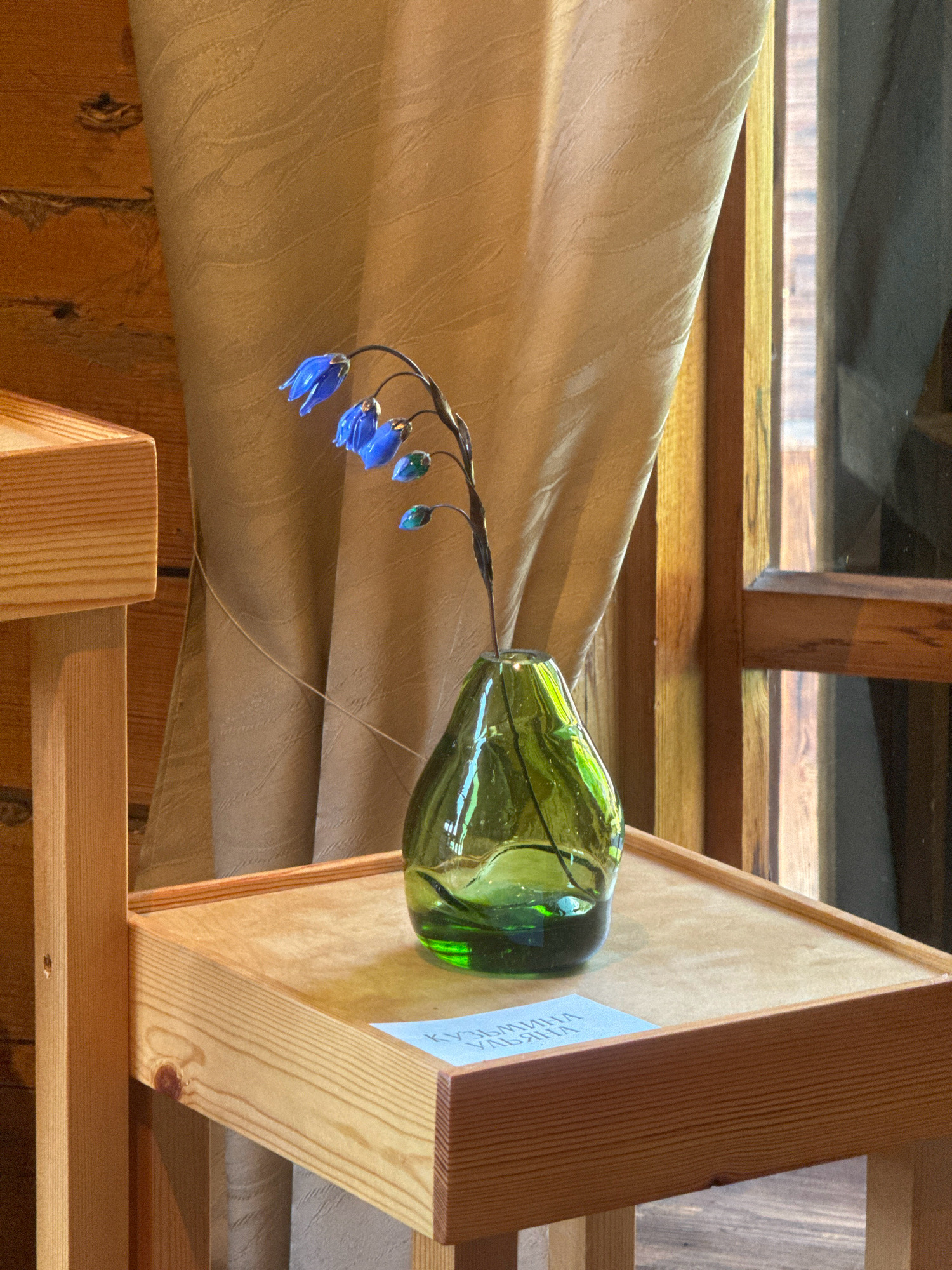 A glass vase containing a decorative glass sculpture of blue flowers with green accents is placed on a light-colored wooden shelf. The shelf is part of a larger wooden structure, possibly a display stand or part of the architecture within a building. Behind the vase and shelf, a beige textured curtain hangs, with a wooden wall visible to the left. To the right, a wooden-framed window or partition is present, with vertical wooden elements visible. A small, light blue card with white text in Cyrillic script is positioned beneath the vase on the shelf. The overall setting appears to be indoors, likely in a museum or cultural exhibition space within the city of Taltsy, Irkutsk Oblast, Russia, given the context of the provided location. No people are visible in the image.