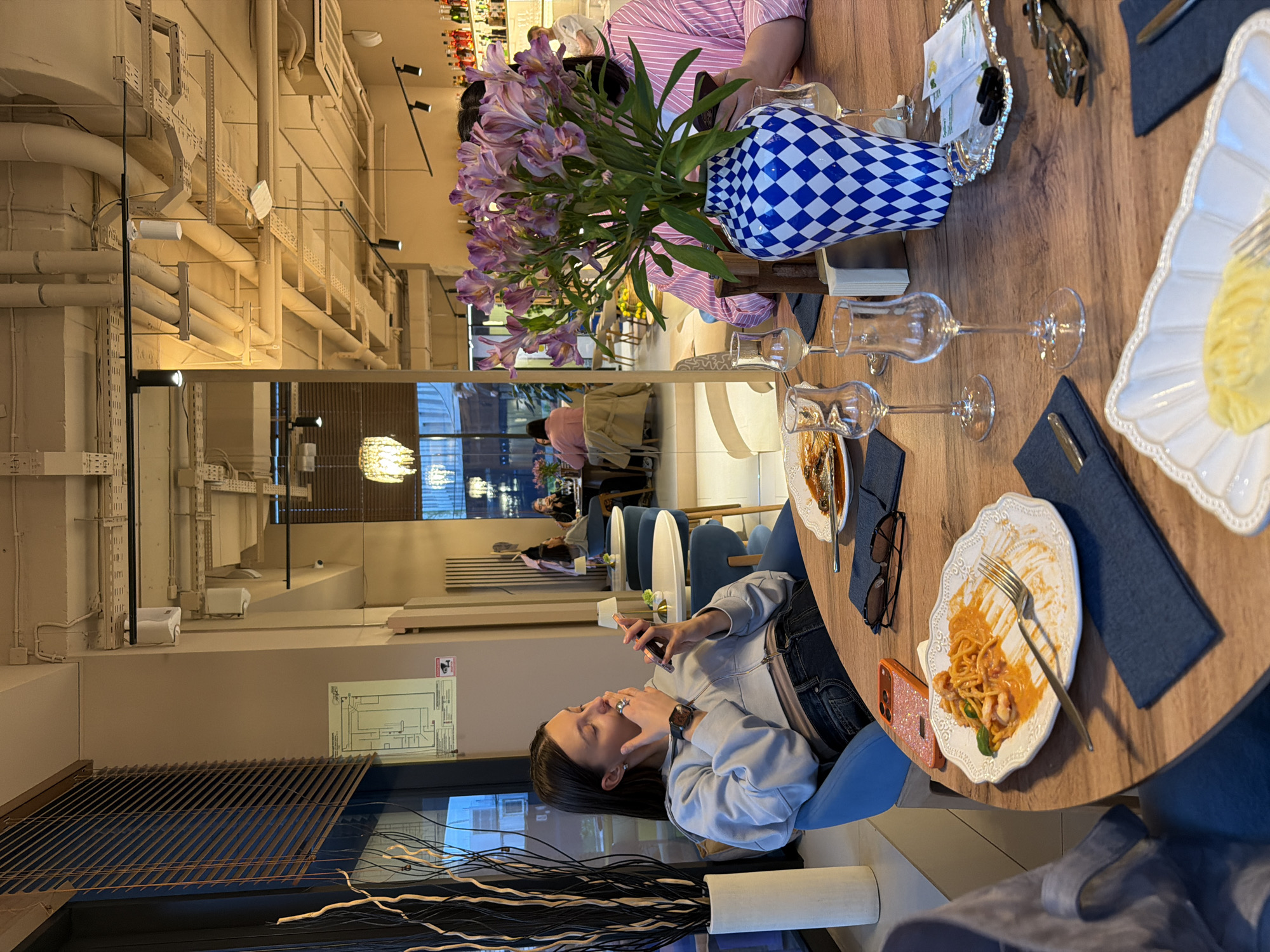A woman in a light blue jacket sits at a round wooden table in a restaurant in Khimki, Russia, using her smartphone. The table is set with plates of pasta, wine glasses, and blue napkins. A blue and white vase filled with purple alstroemeria flowers sits in the center of the table, partially obscuring another person across the table wearing a pink striped shirt. Cutlery, sunglasses, and small packages of sugar are also on the table. The restaurant setting includes a large window with decorative tree branches, a mirrored wall with visible ductwork and lighting fixtures, and other patrons dining in the background.