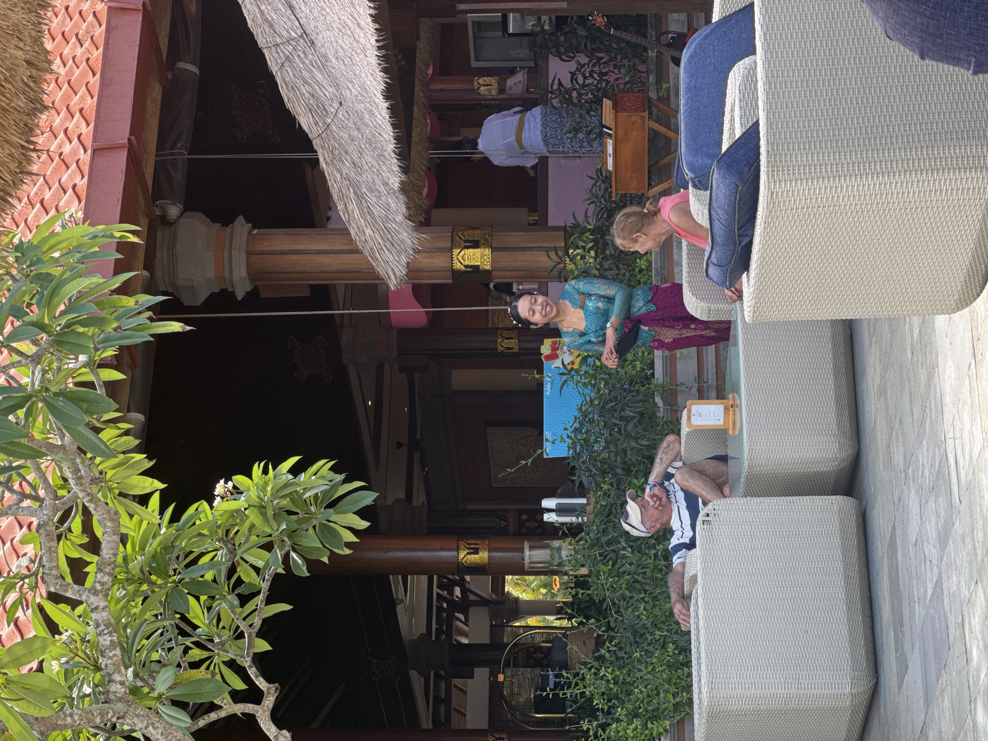 The image depicts an outdoor seating area at a resort in Kuta Selatan, Indonesia. In the foreground, a man in a striped shirt and shorts sits in a white woven armchair, looking to his right with his hand on his chin. To his right, a young girl with blonde hair in a pink top is seated in a similar armchair. Between them is a low glass-topped table. A woman in a turquoise and magenta traditional outfit stands to the left of the girl, facing the man and holding a dark object. Behind them, a thatched-roof structure with wooden pillars and ornate gold detailing is visible, with another person in a white uniform visible in the background. Lush green foliage surrounds the seating area. The overall scene suggests a relaxed, tropical resort setting.