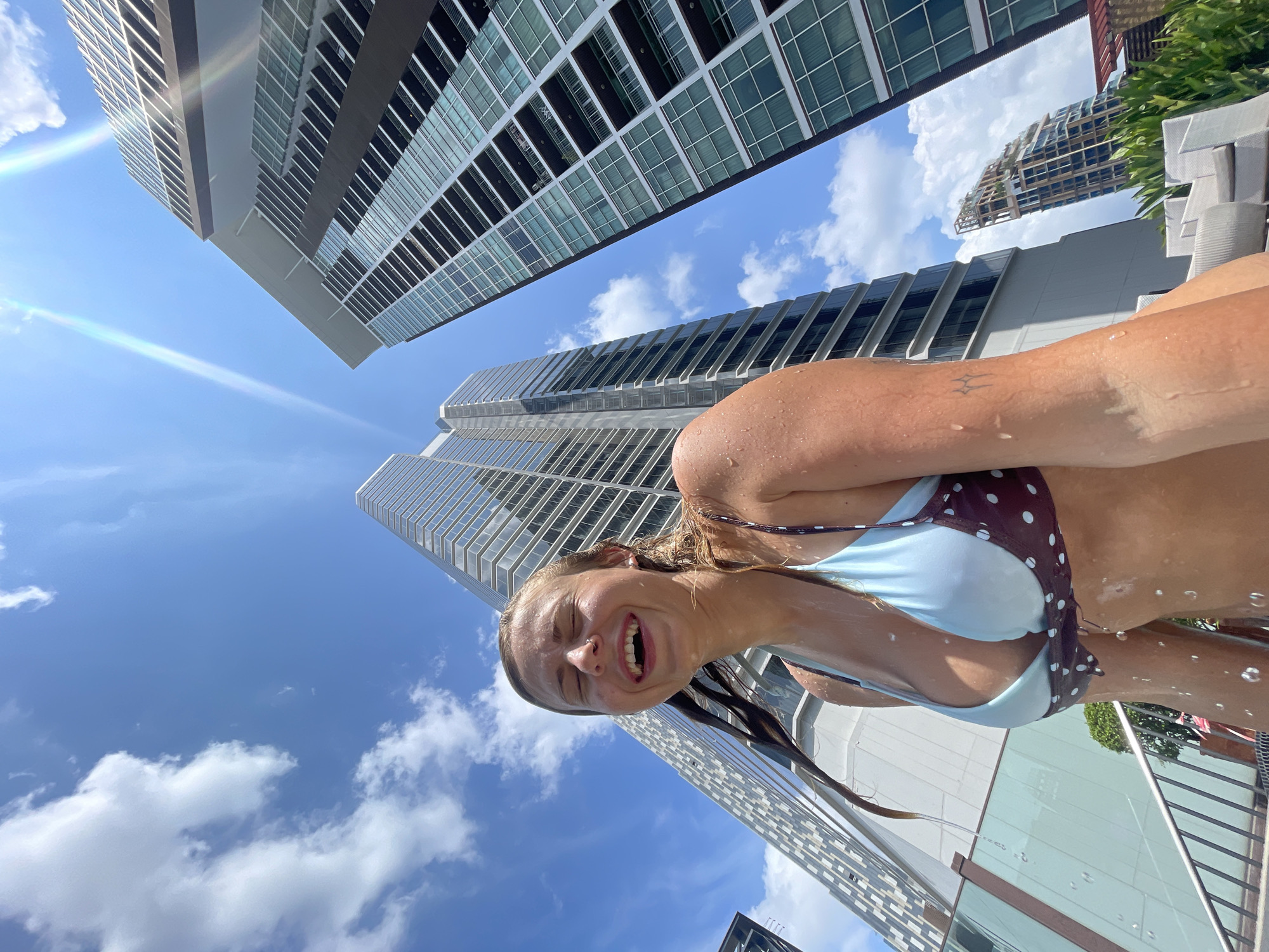 A woman in a blue and brown polka-dot bikini is photographed from a low angle, with skyscrapers and a blue sky with scattered clouds in the background. She is wet, with water droplets visible on her skin, and is smiling broadly with her eyes nearly closed. The location appears to be a rooftop pool area in Bangkok, Thailand, judging by the architectural style of the buildings and the tropical climate suggested by the sky. The photograph appears to be taken with a wide-angle lens, causing a distortion of the perspective. There are building structures and what seem to be trees to the right side of the photograph.