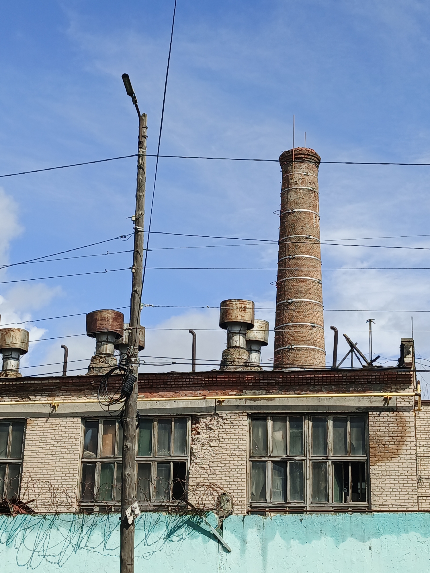 A low-angle, medium shot captures the facade of a weathered industrial building against a bright blue sky with scattered clouds. The building is constructed of light-colored brick with a darker brick cornice. A prominent, tall, red-brick smokestack with visible metal bands rises from the roof, slightly to the right of the center. To the left of the smokestack, on the roof, are several large, circular metal vents with rusty exteriors. A wooden utility pole stands in the foreground, running vertically through the frame, covered in a tangle of black electrical wires and conduits. Barbed wire is visible along the lower section of the building's facade. The windows of the building are large and divided into many panes, appearing grimy and some are broken or covered with makeshift materials. A light blue wall forms the base of the building, with shadows of the barbed wire cast upon it. The image appears to be taken in Zlatoust, Russia, given the context. There are no people or animals visible in the frame, and no discernable actions or interactions are taking place. The scene conveys a sense of disrepair and abandonment.