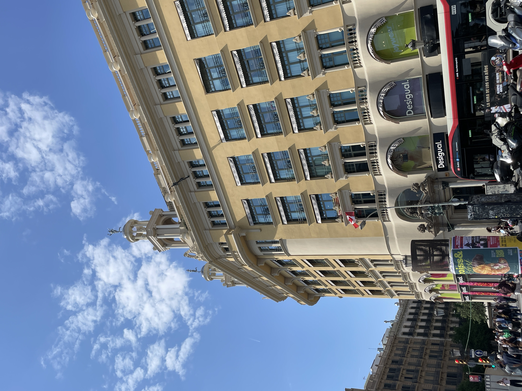 This image captures a vibrant street scene in Barcelona, Spain, dominated by a large, ornate yellow building under a clear blue sky with scattered white clouds. The building, featuring multiple floors with numerous windows, showcases classical architectural details including decorative balustrades, columns, and a small, elegant tower with statues on its rooftop.
On the ground level of the yellow building, several retail stores are visible with large arched windows and signage. Prominently displayed are multiple "Desigual" logos and a "joywear" sign. A red and black articulated city bus, marked "AMB Bus Metropolità," occupies a significant portion of the street, with motorcycles and scooters parked nearby. A crowd of people can be seen walking on the pavement and crossing the street, indicating a bustling urban environment. Traffic lights are also visible, suggesting an active intersection.
Notable details include the bright daylight, suggesting a sunny day, and the detailed architecture of the main building. A flag, possibly Catalan or Spanish, is visible hanging from a balcony on the yellow building. In the background, another, darker building adds depth to the urban landscape.
Visible text includes multiple instances of "Desigual" and "joywear." On the bus, "AMB Bus Metropolità" is clearly legible. Several advertisements are present, including one for "LA GIRA DE LOS 100 AÑOS" featuring "OLIMPIO 23 MAYO" at "ARENA BARCELONA." Another partially visible poster contains text like "qual. Fyval. Egual. YOUSS& Fest DEL DISC BE." Numbers such as "437" and "190" are also discernible, possibly relating to bus routes or street addresses. The overall scene depicts typical daily life in a lively European city.