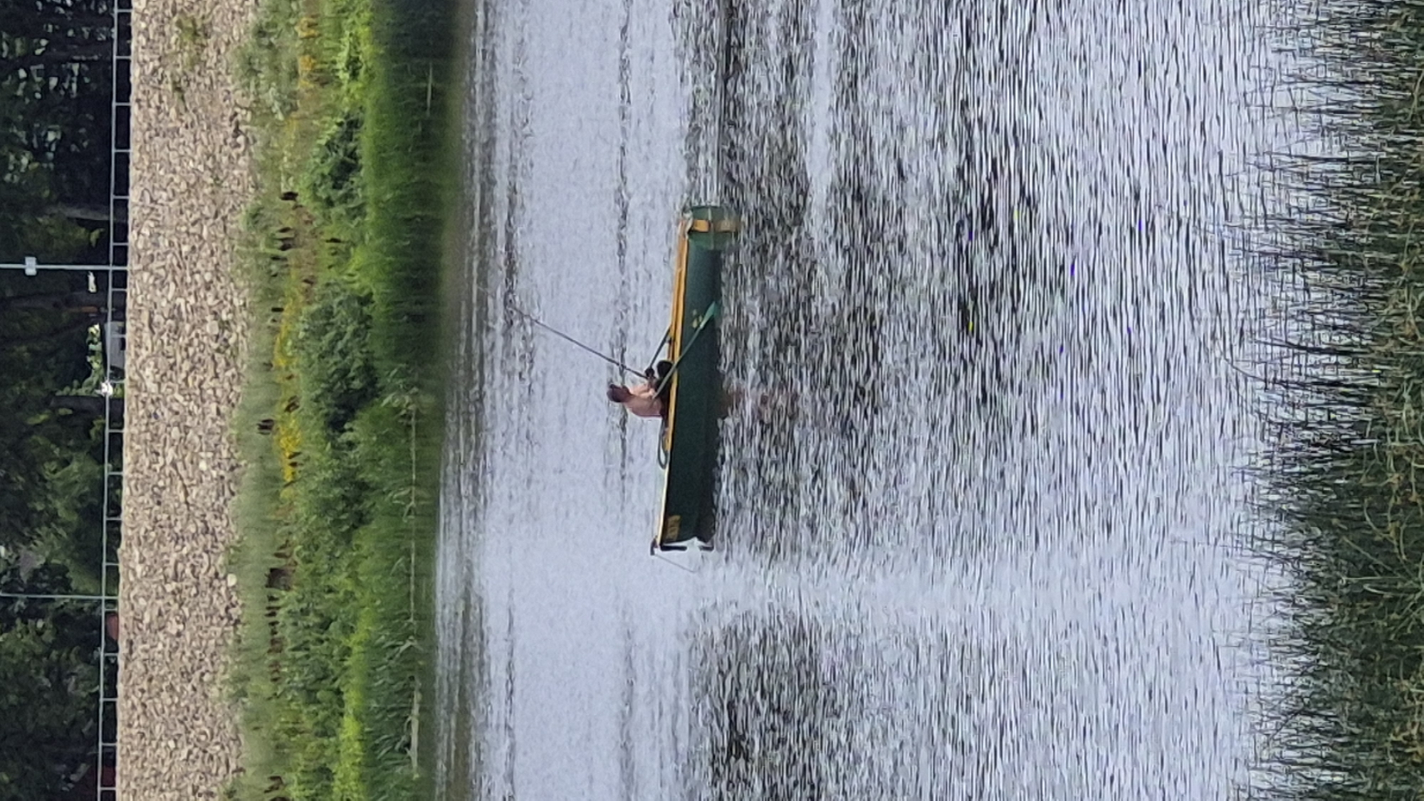 An unidentified person is fishing from a small, green and yellow boat on the Daugava River in Jēkabpils, Latvia. The person is seated and holding a fishing rod. The river has a rippled surface reflecting the overcast sky. In the background, a steep, rocky embankment is visible, covered with green vegetation. Reeds and tall grass line the foreground of the river. The image appears to have been taken from a slightly elevated position.