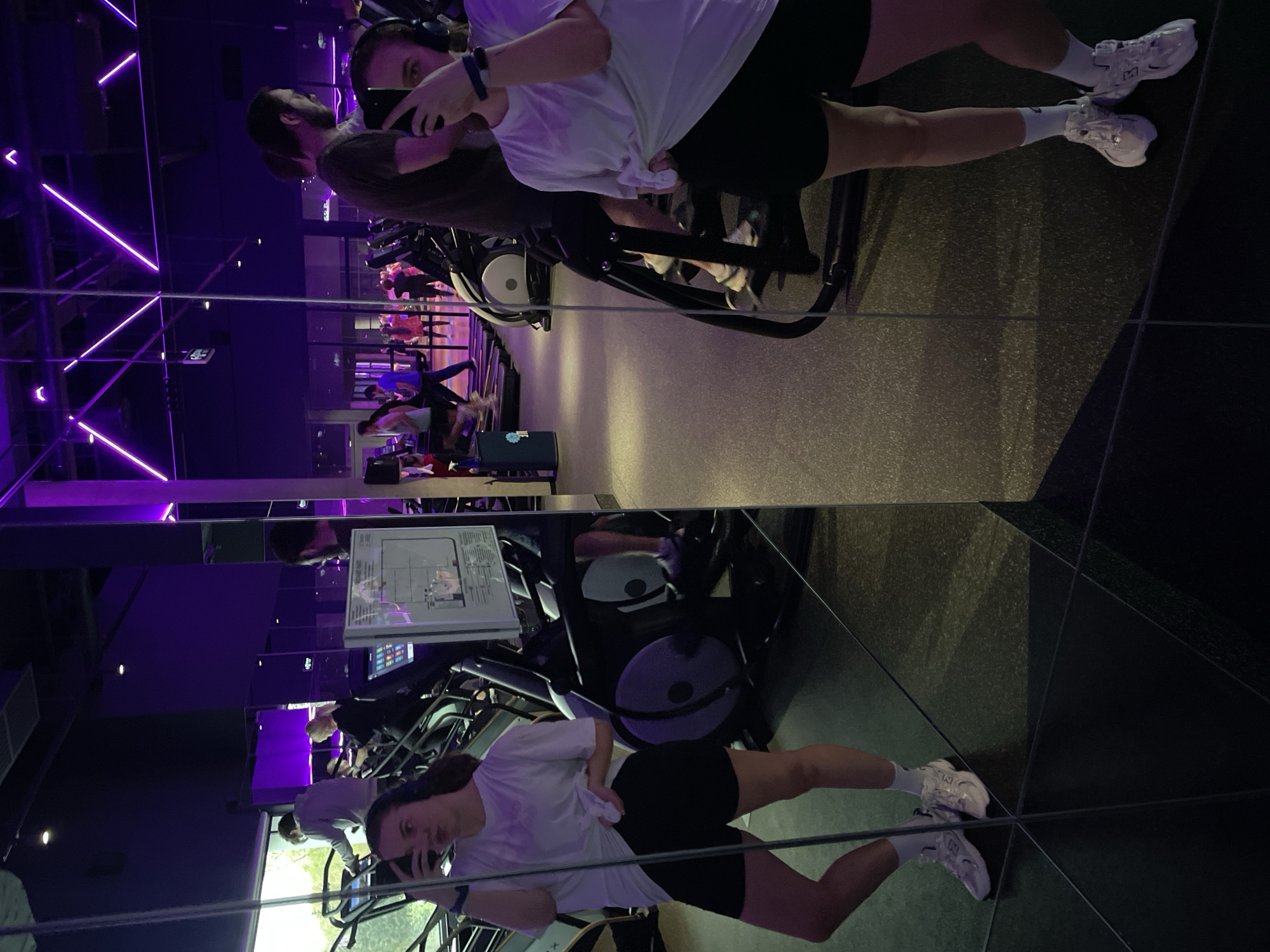 An indoor scene captured in Moscow, Russia, shows a young woman taking a mirror selfie in what appears to be a modern gym or fitness studio. The image is dominated by the reflection in large mirror panels that cover the walls, creating a layered and somewhat disorienting perspective.
The main subject, positioned towards the center-right of the frame (when upright), is seen from the chest down in the mirror, holding a dark smartphone to capture her reflection. She is wearing a white t-shirt, black athletic shorts, white socks, and white "New Balance" sneakers. A smartwatch is visible on her left wrist, and headphones are draped around her neck.
In the reflections, several other individuals are visible, actively exercising on various gym equipment such as treadmills and ellipticals. The setting features dark walls and floors, contrasted by striking purple neon strip lights running across the ceiling and walls, giving the space a dynamic and contemporary ambiance. Some additional spotlights are also visible on the ceiling. A detailed informational sign or poster, though illegible, is reflected on a wall in the background. The distinct reflections and strategic lighting suggest an evening or night-time setting. The overall mood is focused and active, typical of a workout environment.