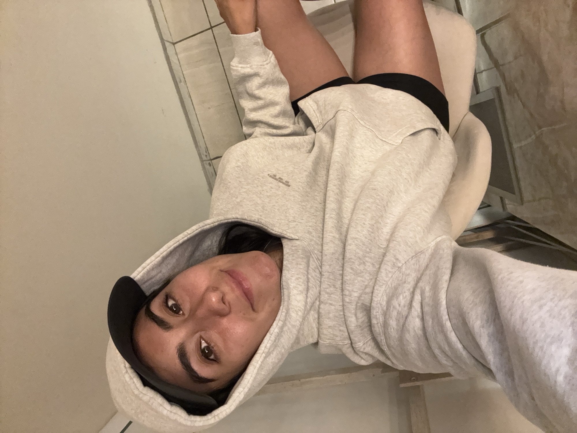 The image shows an individual, identified as a young adult female, seated indoors. She is oriented slightly upwards relative to the camera, indicating a self-portrait. The subject wears a light grey hooded sweatshirt with the hood pulled over her head, partially obscuring a black baseball cap. A small, light-colored embroidered logo or text is visible on the upper left sleeve of the sweatshirt. Dark shorts are visible beneath the sweatshirt on her right thigh. She possesses dark hair, dark eyes, and a slight smile while looking directly at the camera. Her right arm is visible, bent at the elbow. A light-colored chair or seat back is visible behind her. The background on the right features light-colored square tiles with dark grout lines, transitioning to a plain, light-toned wall or ceiling. A plain, light-colored vertical surface is present on the left. The scene is evenly lit by artificial light. No distinct visual cues are present within the image to specify a particular location within the city of Puebla, Mexico.