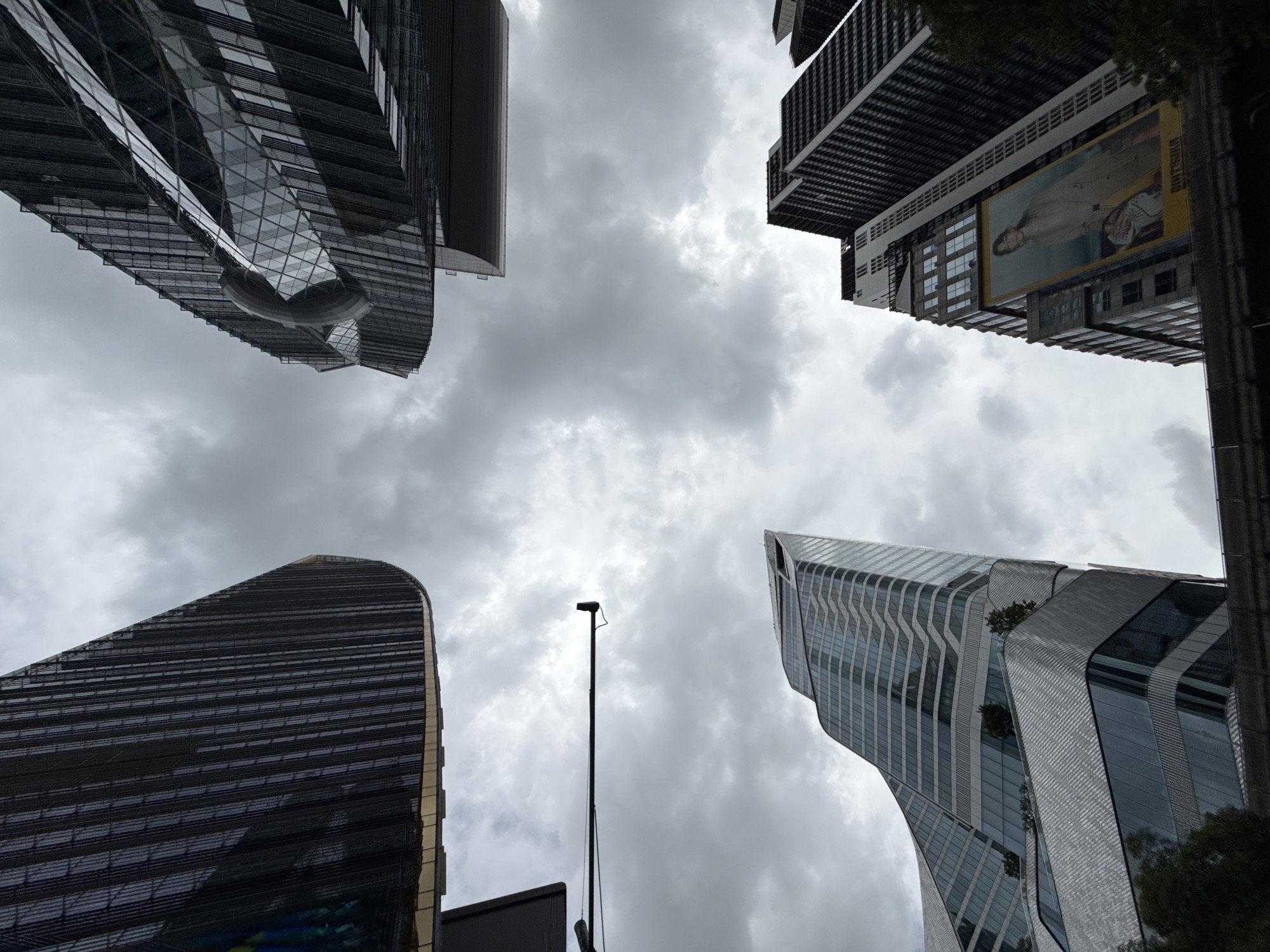 This is a low-angle shot of several modern skyscrapers in Bangkok, Thailand. The sky is overcast with grey clouds. The buildings are constructed with glass and metal, featuring sharp angles and curved surfaces. One building has a prominent advertisement featuring a woman wearing a trench coat. There is a street light pole visible in the upper left portion of the image. The image captures an urban landscape with a focus on architectural design.