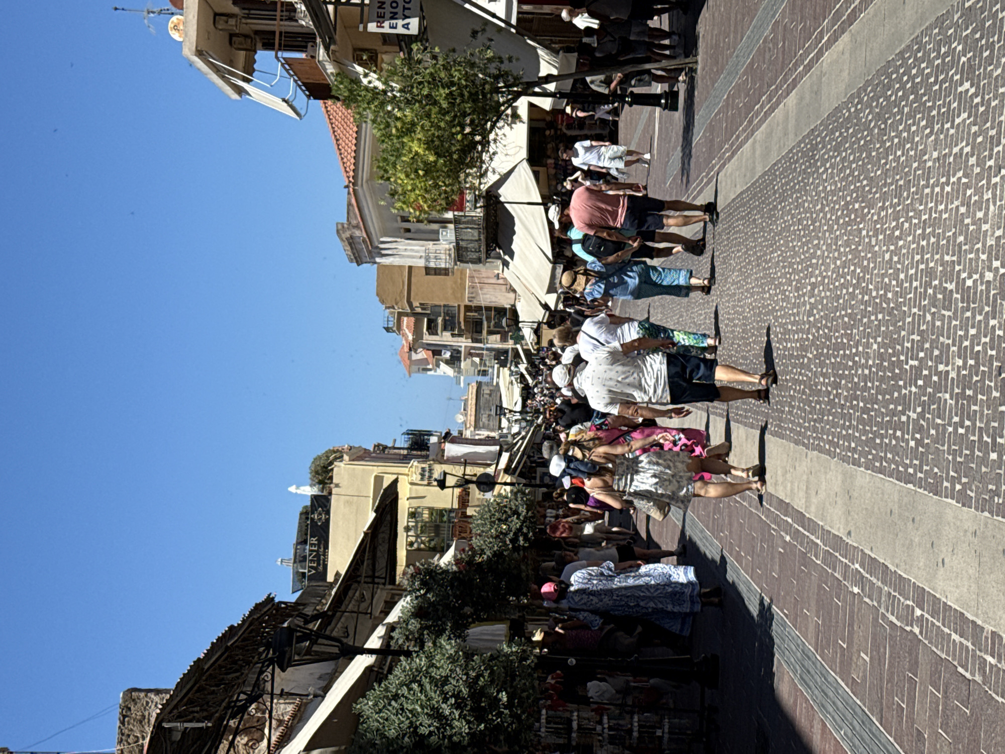 The image depicts a crowded pedestrian street in Chania, Greece, likely during daylight hours due to the bright sunlight casting distinct shadows. The street is paved with cobblestones and brickwork, suggesting a historic area. On either side of the street are buildings with shops and awnings, some displaying signage. Several trees are planted along the walkway, providing a touch of greenery. A diverse group of people, dressed in casual summer clothing, are walking in both directions, some browsing shop displays. The sky is a clear, bright blue. The scene is characteristic of a busy tourist area, possibly within the Old Town of Chania, given the architecture and paving style.