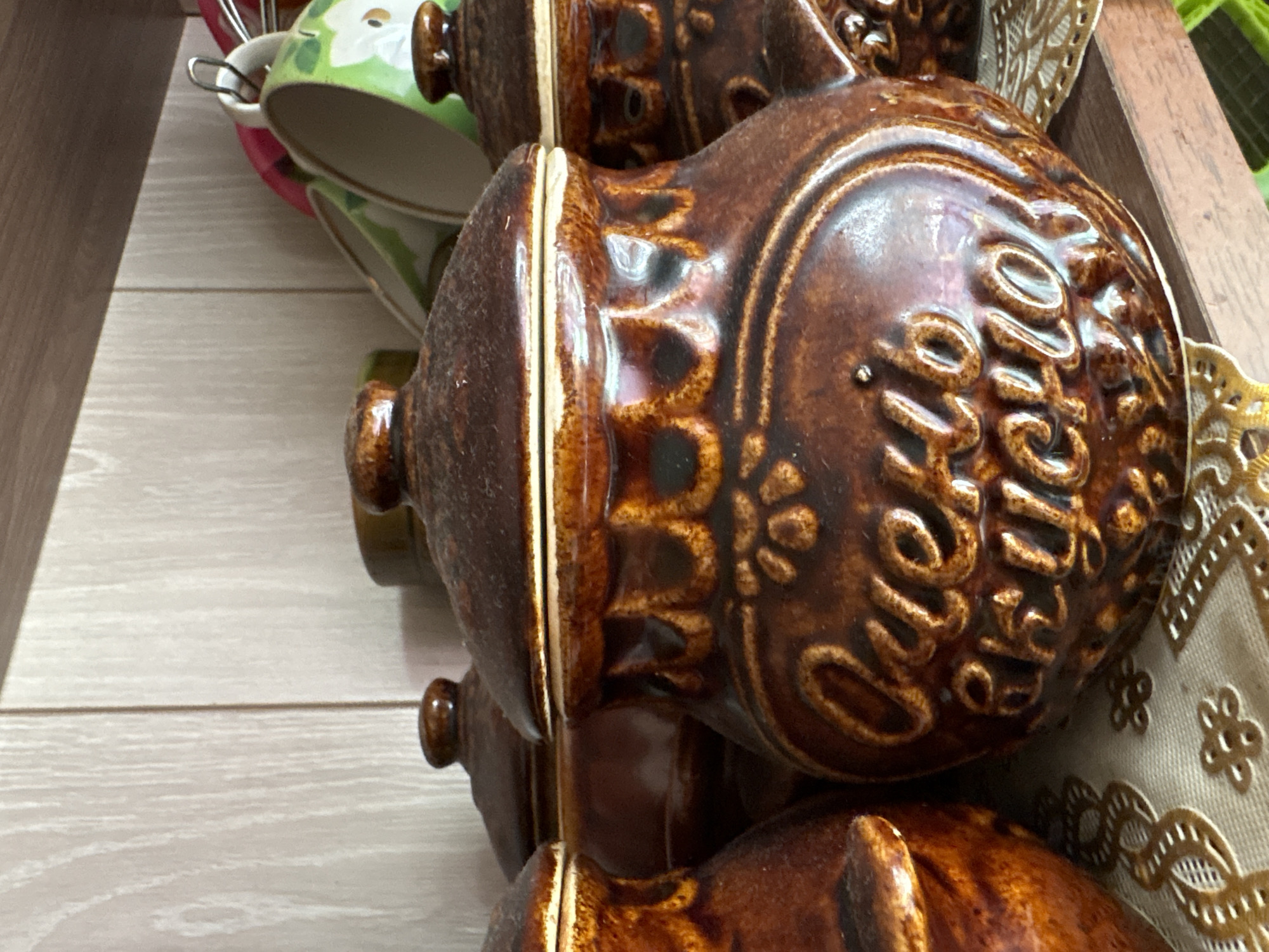 A close-up, eye-level shot shows a collection of brown ceramic jars with lids, arranged on a shelf against a light-colored, wood-paneled background. The jars have embossed decorative patterns and lettering in a lighter brown shade. The jar in the foreground is the most detailed, displaying the Cyrillic text "Очень вкусно" which translates to "Very tasty." The other jars are partially visible, suggesting a set. In the background, on a higher shelf, a rack of colorful mugs, primarily green and pink, is also visible. The lighting is from the front, casting subtle shadows and highlighting the glossy finish of the ceramic. The overall scene appears to be a domestic setting, likely a kitchen or pantry, with no people present. The location in Vetluzhsky, Russia, is inferred from the context of the Cyrillic text and the rustic aesthetic.