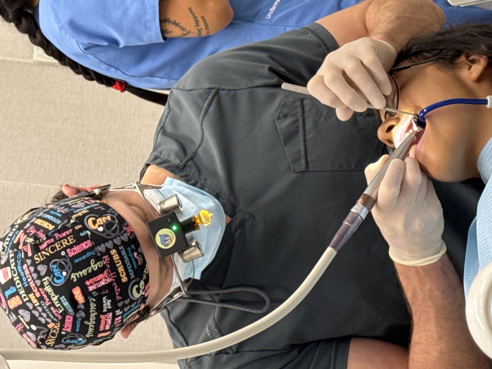 A dentist, wearing a dark scrub top and a surgical cap with various words and heart symbols, is performing a dental procedure on a patient. The dentist is also wearing loupes with a light attached. The patient is lying down, with their mouth open. A dental tool is in the patient's mouth, and another tool is being held by the dentist. A blue suction tube is also visible in the patient's mouth. In the background, another person wearing a blue scrub top with a braided black ponytail is visible. The environment appears to be a dental clinic, with neutral-colored walls. There is no indication of the image being taken in Barcelona, Spain.